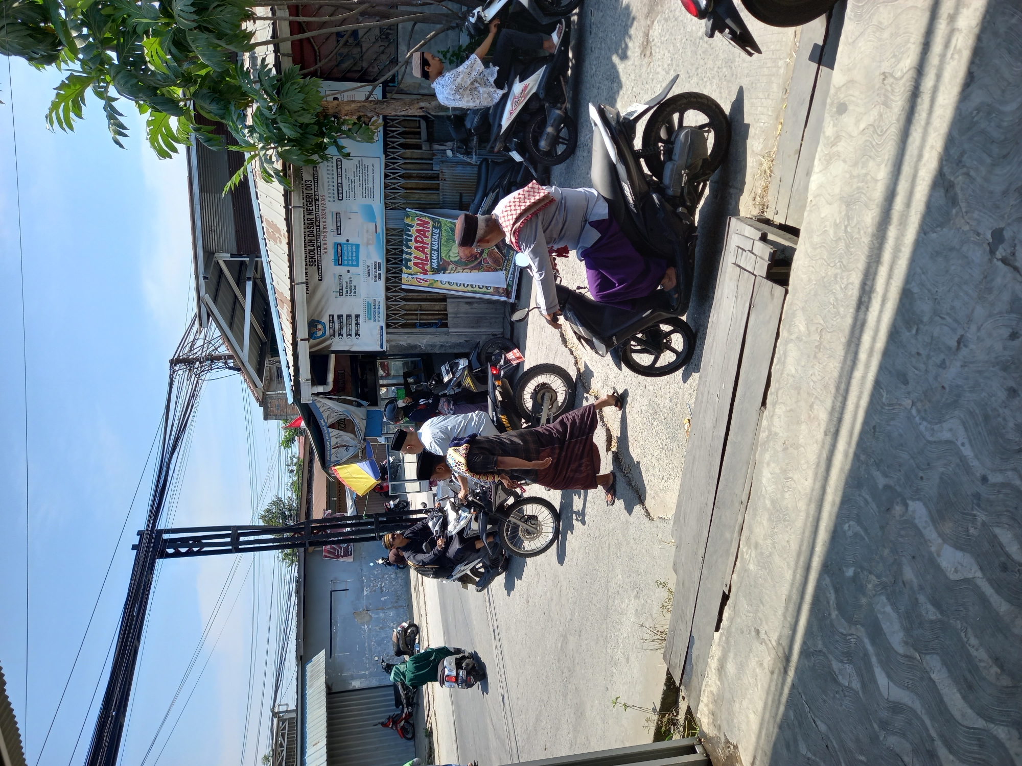 The image depicts a street scene in Samarinda, Indonesia, likely on a sunny day given the strong shadows. In the foreground, a man wearing a peci (cap), a grey shirt, and a purple sarong is riding a black scooter. He has a patterned cloth draped around his shoulders. To his right and slightly behind him, another man in a white patterned shirt and a white peci is also riding a scooter. In the background, several other people are on motorcycles. One man in a green hooded jacket is on a white scooter. Another man wearing a white peci is on a black motorcycle, and a man in a black shirt and white peci is on a scooter. There are buildings visible, including one with a banner that appears to be an advertisement or public notice. The street is paved, and there are power lines overhead. The overall impression is of a typical urban street in Indonesia with people commuting on motorcycles.