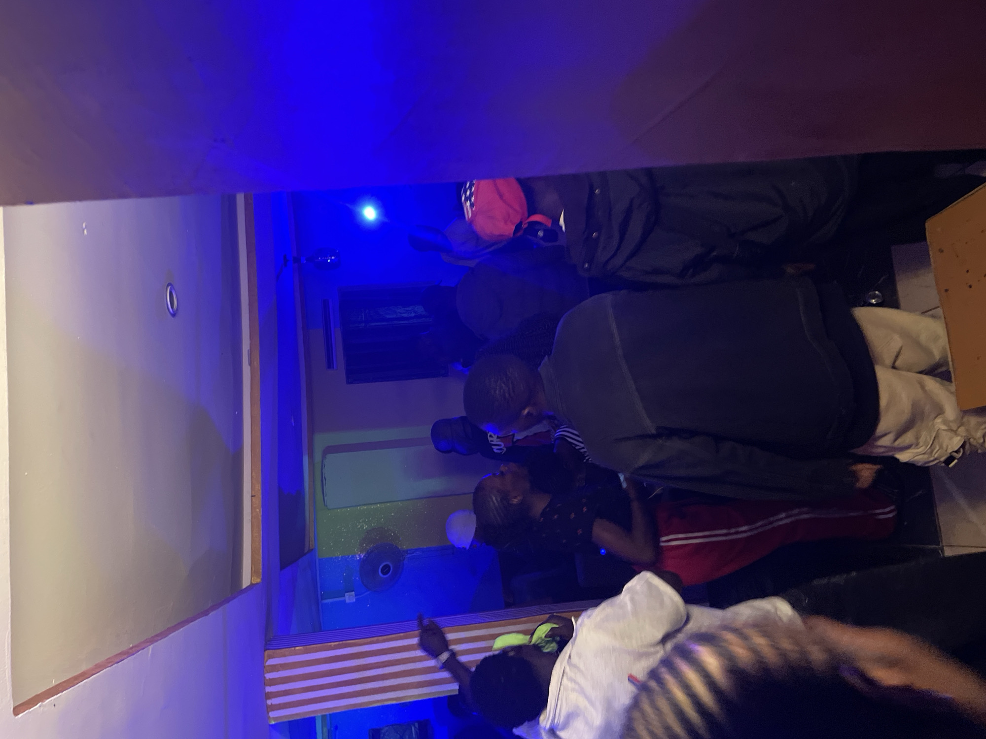 The image depicts an indoor gathering at night, likely a party or social event, in Durumi, Nigeria. The scene is dimly lit, with prominent blue lighting casting a cool, atmospheric glow. In the foreground, a group of individuals are visible, some facing away from the camera, others in profile, engaged in conversation or simply present at the event.
To the left, a person wearing a white shirt is partially visible. Next to them, a woman in red pants with white stripes is interacting with another individual. Behind them, a fan is mounted on a wall. The overall environment appears to be a modest indoor space, possibly a bar, club, or private residence. The blue lighting dominates the scene, creating shadows and highlighting certain areas. The presence of a fan and air conditioning unit suggest a warm climate. The people are dressed casually, indicating a relaxed social setting. The image captures a moment of social interaction within this vibrantly lit, nighttime environment in Durumi.