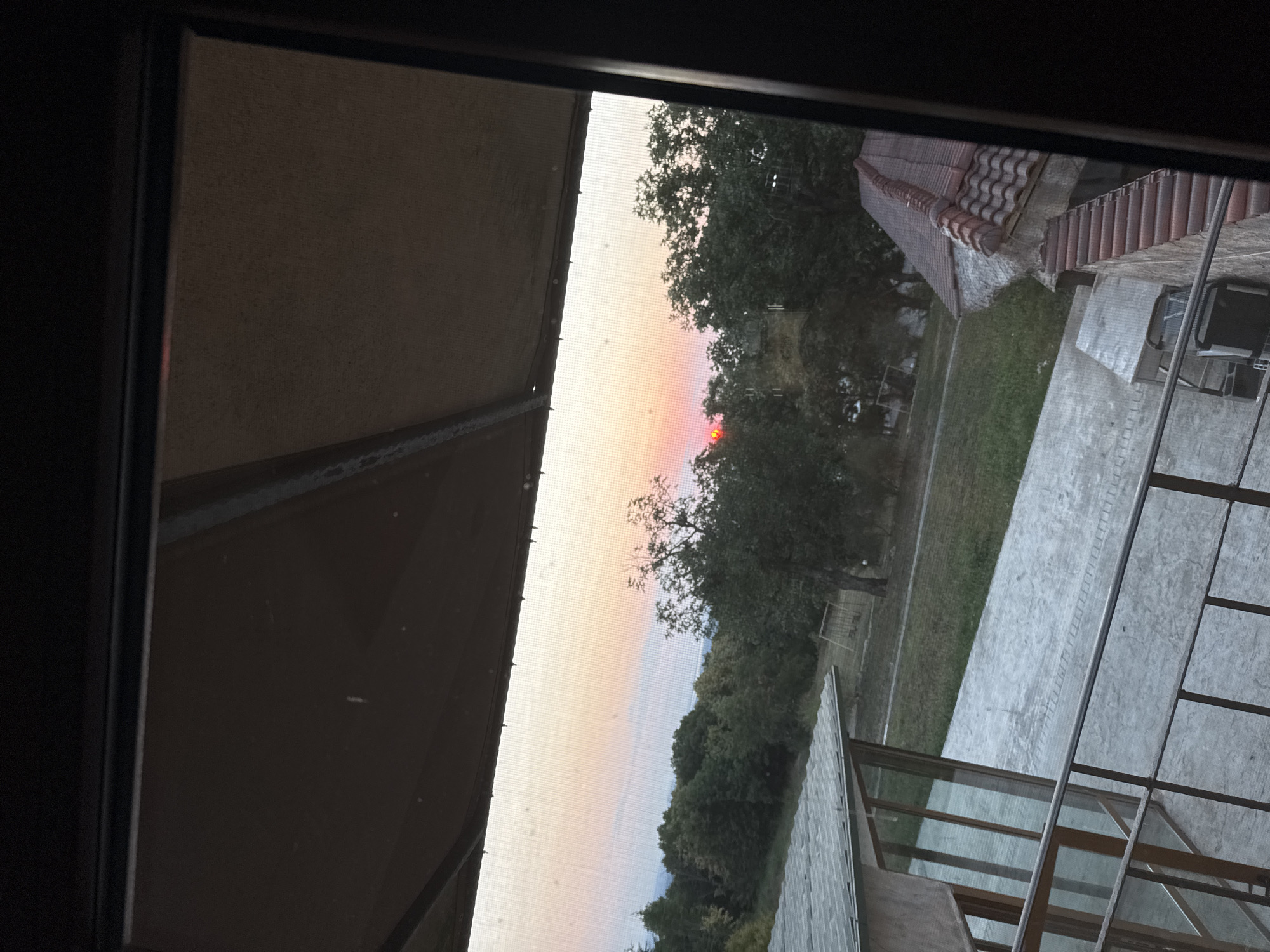 The image shows a sunset over a rural landscape viewed through a screened window. In the foreground, a balcony railing is visible, with part of a building to the left and a tiled roof of another building to the right. The background consists of trees and a gently sloping grassy area. The sky displays a gradient of colors from soft pink and orange near the horizon to a lighter hue above, indicating the setting sun. The overall impression is a peaceful evening scene in a natural setting, possibly a residential area or a rural property in Sredets, Bulgaria.