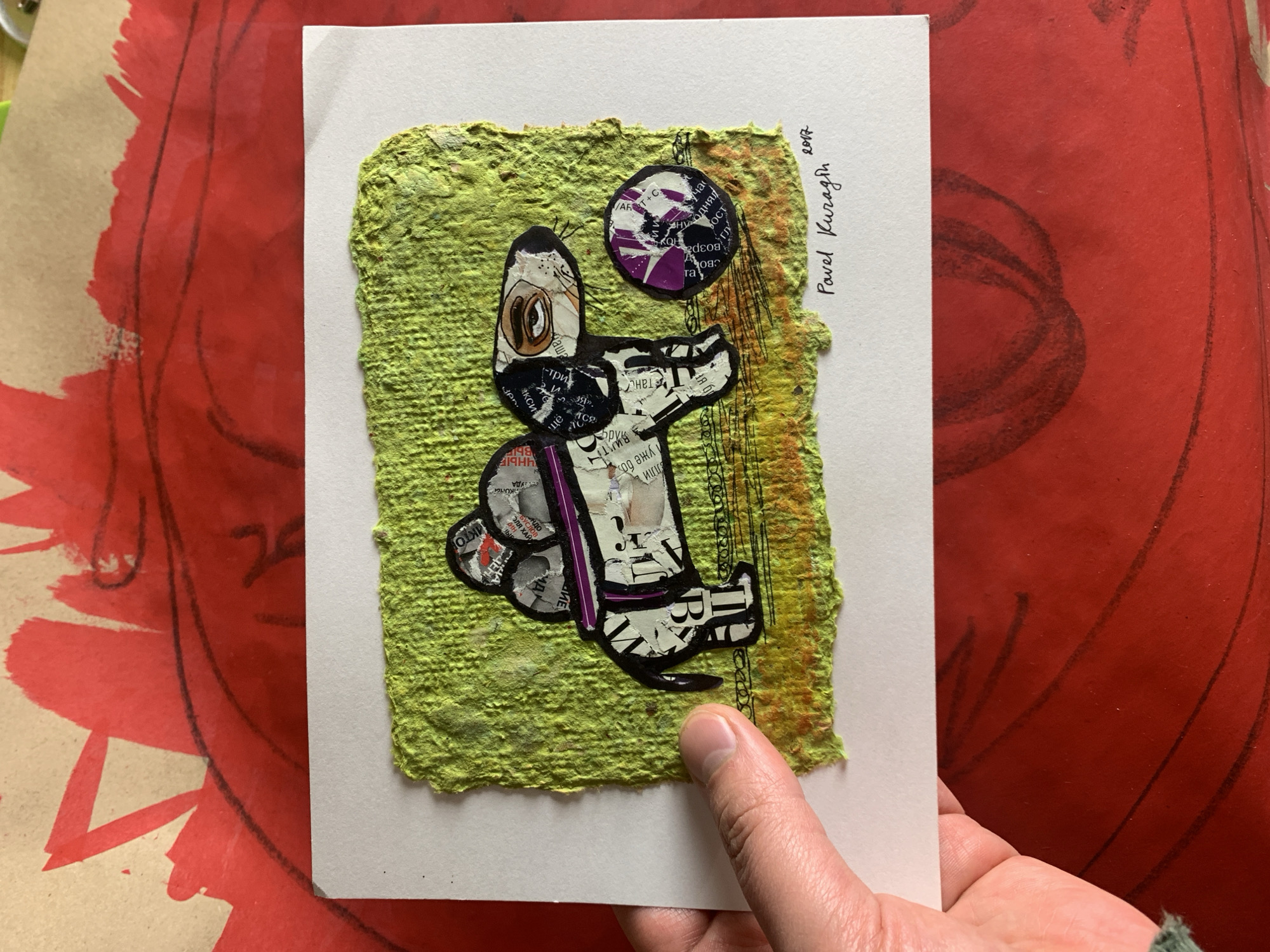 The image shows a close-up of a collage artwork featuring a black and white dog with a purple collar, standing next to a purple and black ball. The dog and ball are made from cut-out pieces of text and images from magazines or newspapers. The artwork is mounted on a textured, rough green paper that is slightly torn at the edges. This green paper is attached to a white card. The white card is placed on top of a larger red surface with abstract black lines and red brushstrokes. The artwork is signed "Pavel Kuragin 2017" in the lower right corner. A person's thumb and forefinger are visible on the left side of the white card, holding it in place. The artwork is likely displayed in an indoor setting, given the lighting and the presence of the red background which appears to be another piece of art or material. No specific details about the city of Sergeevo, Russia, are discernible from the image itself; this information would be external context.