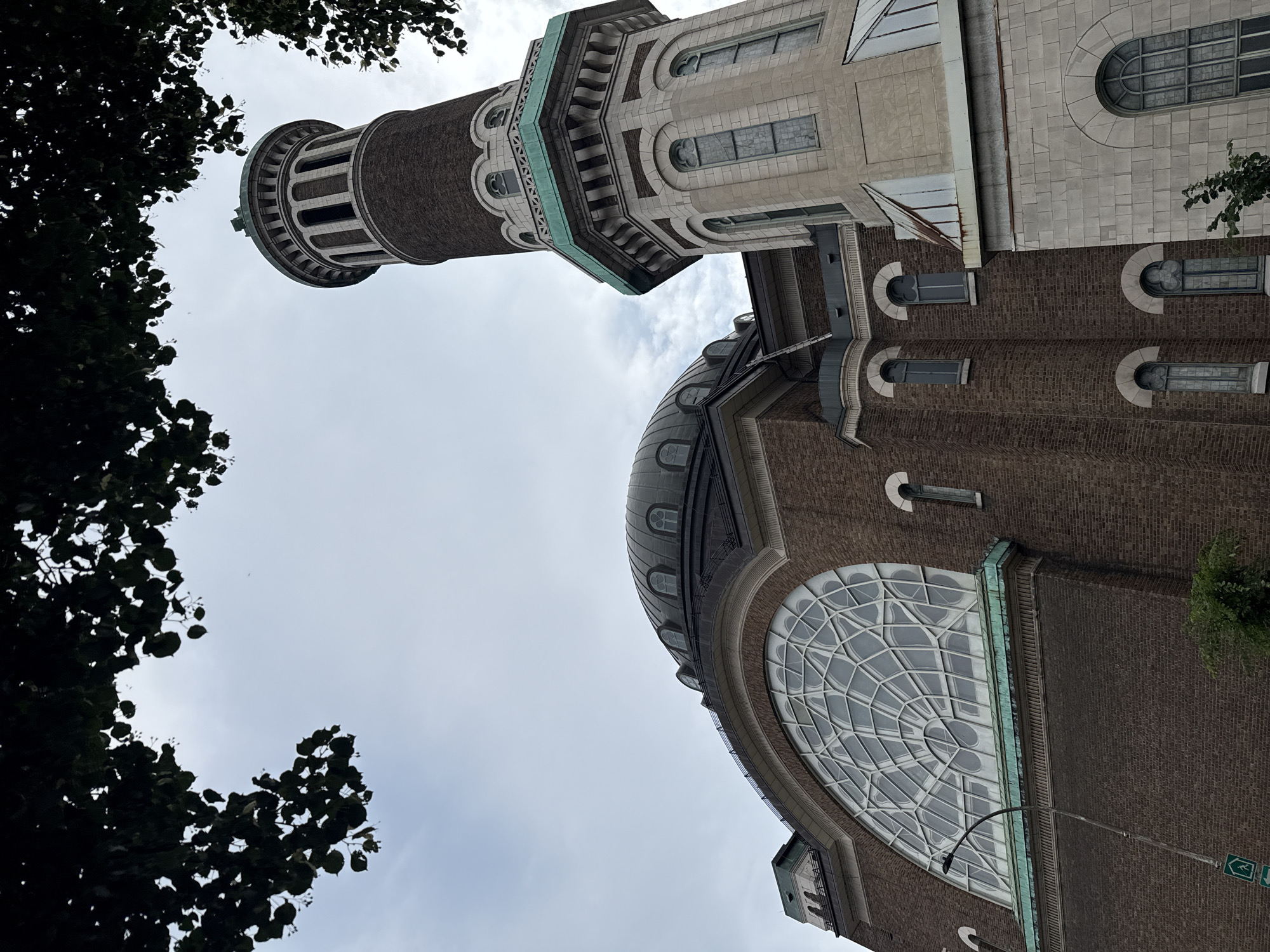 This image shows the exterior of a large brick building with a prominent domed roof and a tall, cylindrical tower. The building's architecture features arched windows, decorative cornices, and a large, semi-circular window with intricate latticework on the main facade. The tower is clad in stone at its base and brick higher up, with a green-patinated cap. Green foliage from overhead trees frames the top of the image, suggesting an outdoor setting. The sky is overcast. This building is the Congregation Shaar Hashomayim synagogue, located at 4255 Kensington Avenue in Montreal, Canada. There are no people visible in the image.
