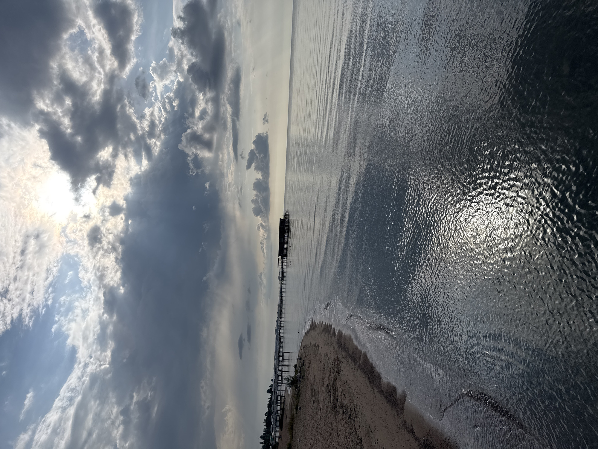 The image depicts a serene lakefront scene in Sary-Oy, Kyrgyzstan. A sandy beach curves along the left side of the frame, meeting the gentle lapping waves of a calm body of water. In the distance, a long wooden pier stretches out into the lake, lined with several small structures or gazebos, each topped with a white, conical roof. Further along the pier, a darker, more substantial building with a dark roof stands, possibly a boathouse or recreational facility. The sky above is dramatic, with a mix of dark, heavy clouds and patches of bright sunlight breaking through, creating a dynamic play of light and shadow on the water's surface. The overall atmosphere is tranquil and picturesque, suggesting a peaceful day at the lake.