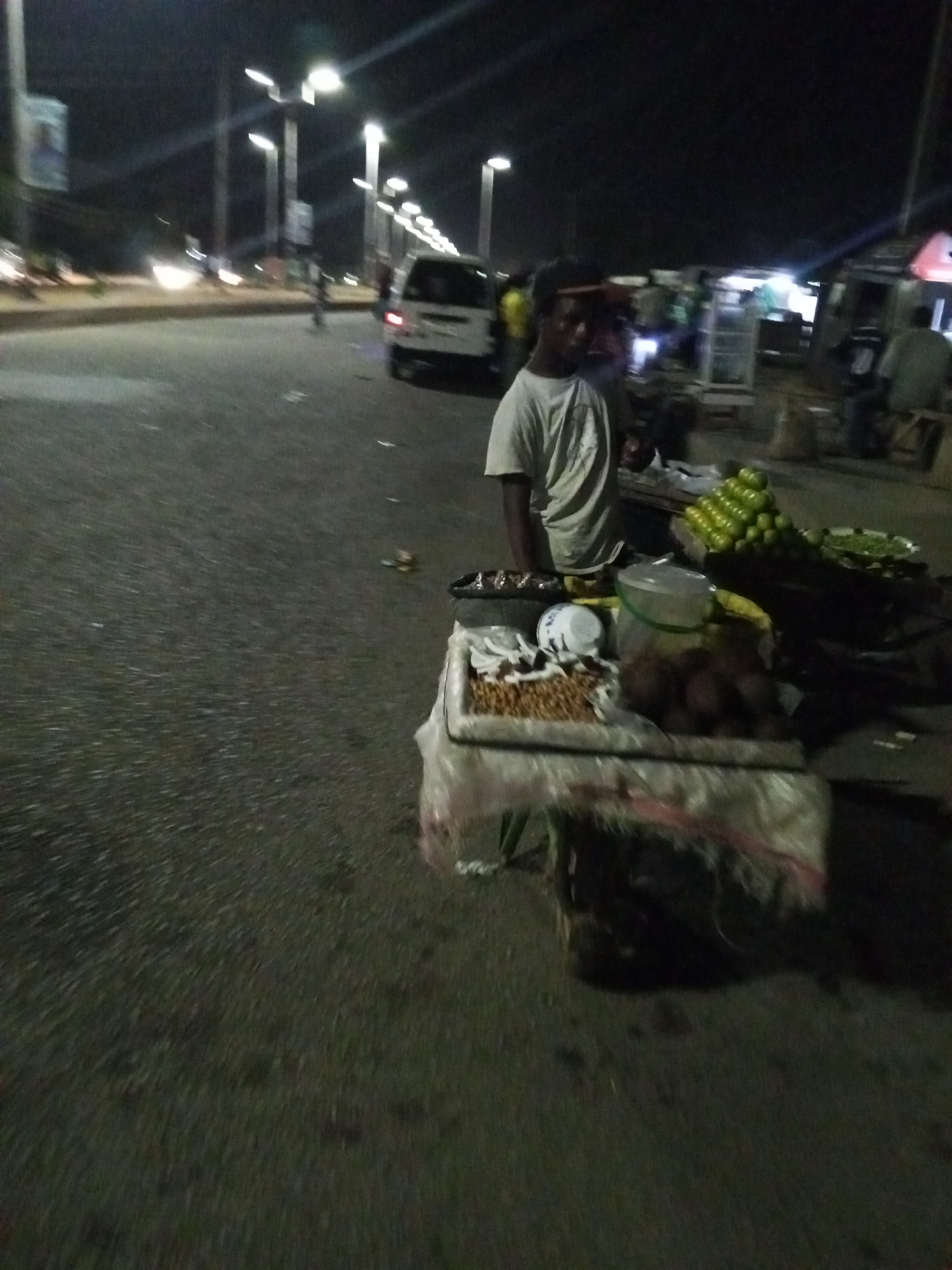 A street vendor stands behind a cart of food under streetlights in what is likely the city of Bukuru, Nigeria. The cart holds green citrus fruits, brown coconuts, white coconut pieces, and other assorted goods. The vendor, who is Black, wears a light-colored t-shirt and a dark cap. A white van passes on the road nearby. Other people and structures are visible in the background. The road surface is dark and appears to be asphalt. The lighting is generally dim, with bright spots from the streetlights.