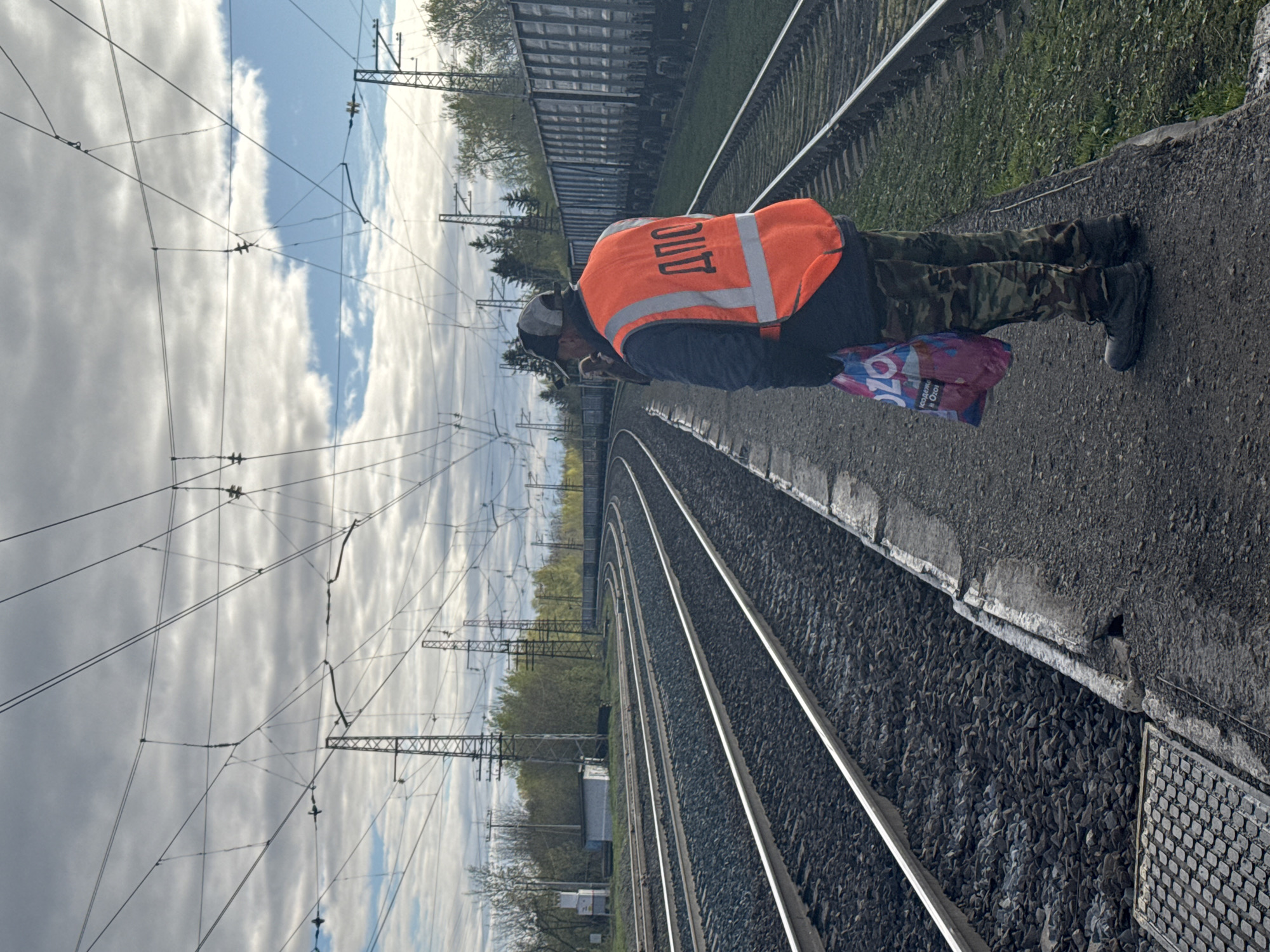 A railway worker wearing an orange safety vest with Cyrillic lettering (ДПС) stands beside a train track. He is also wearing camouflage pants and holding a shopping bag. The train tracks curve into the distance under a cloudy sky. The location is likely Бородулино, Russia. The worker appears to be inspecting the tracks or waiting for a train. Overhead power lines are visible. A railcar is situated on the far-right edge of the frame.
