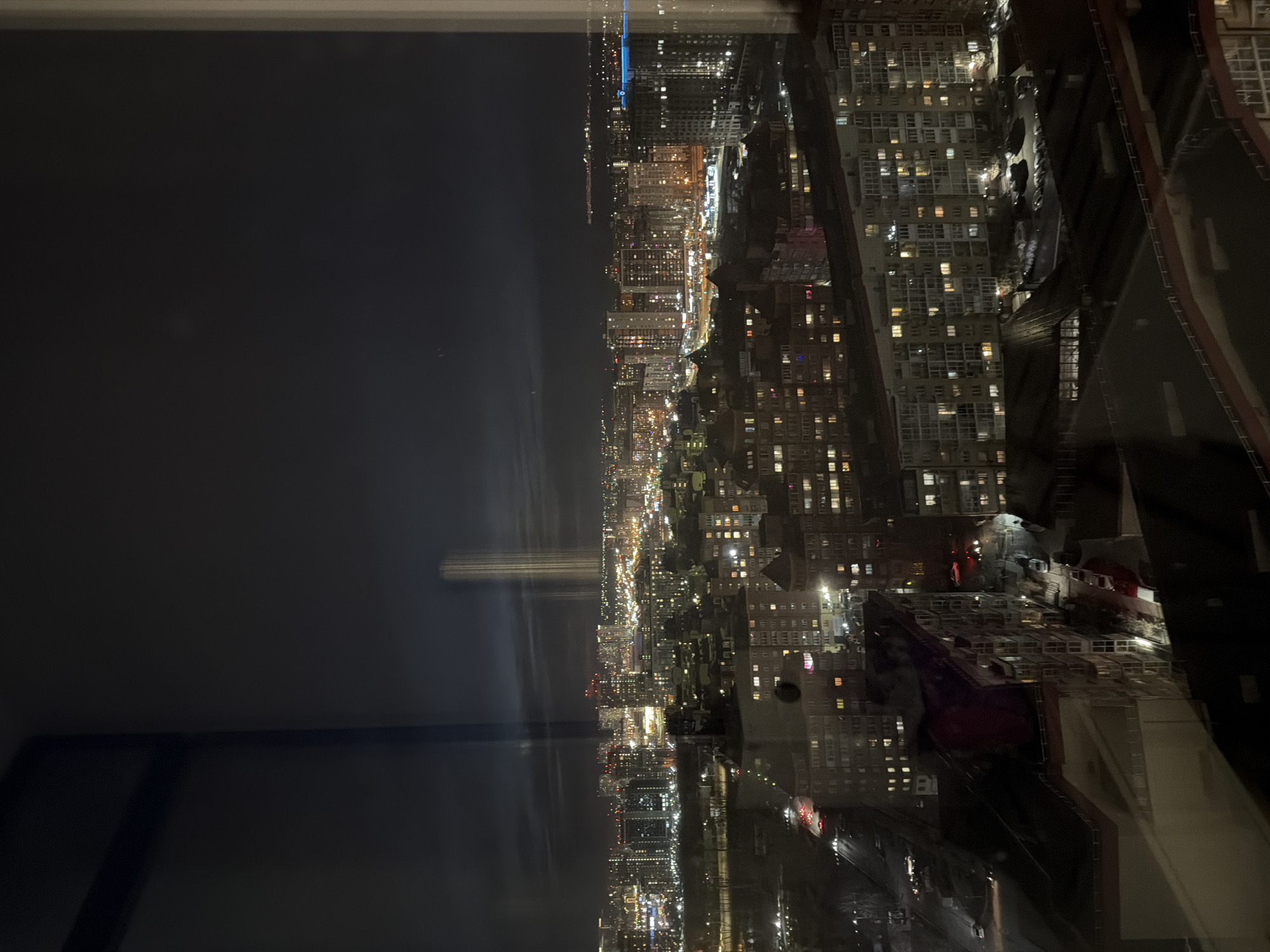 This media file captures a panoramic night view of Krasnoyarsk, Russia, from a high vantage point, likely through a window, given the significant reflections. The cityscape sprawls across the horizon under a dark, clear night sky, showcasing a dense urban environment illuminated by countless lights.
Numerous high-rise and mid-rise buildings dominate the scene, their windows glowing with various hues, indicating an active city at night. The architecture appears diverse, with blocks of apartments and commercial structures densely packed. In the immediate foreground and to the right, reflections on the window glass obscure some details but reveal dark shapes from the interior space.
A prominent feature is a vast, dark expanse, likely the Yenisei River, extending across the middle ground. On the opposite bank of this body of water, a tall, vertical beam of light stands out, possibly an illuminated landmark or monument, reflecting faintly on the water's surface. Further along the distant bank, the city continues to stretch, a vibrant tapestry of residential and street lights, with a distinct bright blue light visible further in the distance.
Lines of streetlights and vehicle lights delineate roads and elevated structures, hinting at the city's infrastructure winding through the buildings. The overall activity is that of a bustling metropolis continuing through the night, with human presence implied by the myriad lit windows. The clear weather ensures excellent visibility of the distant city lights. There is no visible text in the image, and no specific events or people are discernible. The scene conveys the immense scale and vibrant, yet calm, atmosphere of a large city after dark.