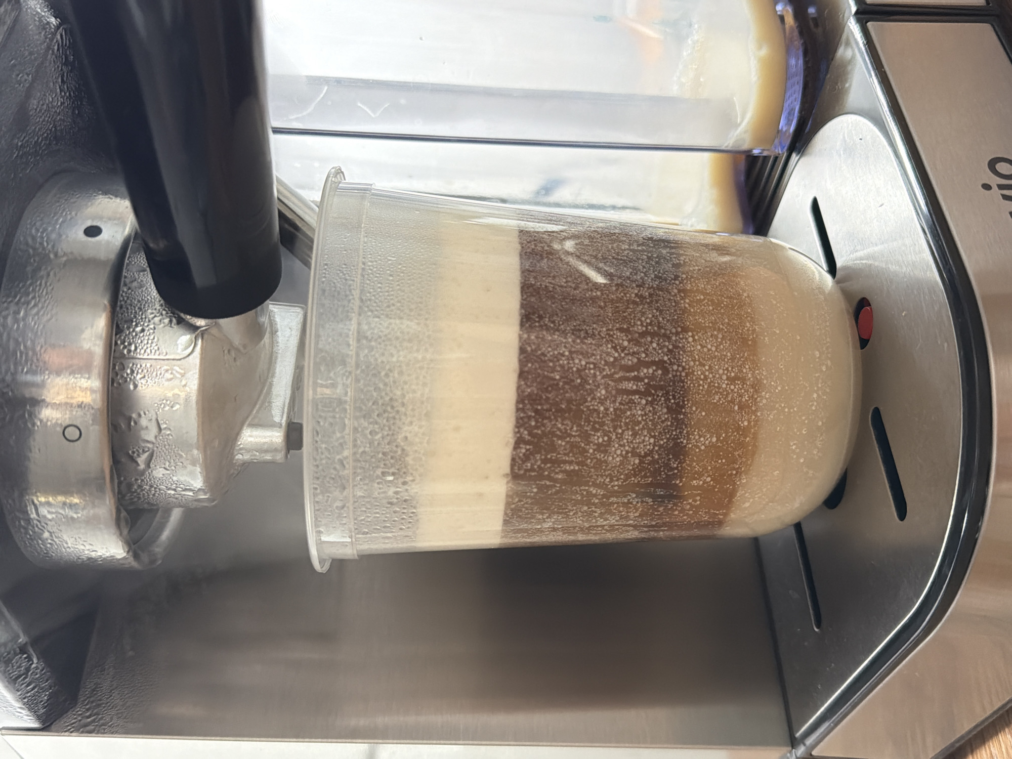 The image depicts a close-up view of a machine dispensing a layered beverage, likely a coffee-based drink, into a clear plastic cup. The machine, partially visible, is stainless steel and features a black dispensing spout. Condensation is visible on the machine's components and the exterior of the cup. The beverage consists of distinct layers of varying shades of brown and beige. The surrounding environment appears to be a clean, stainless steel surface. The location in Reshetnikovo, Russia, is not ascertainable from the visual content of the image.
