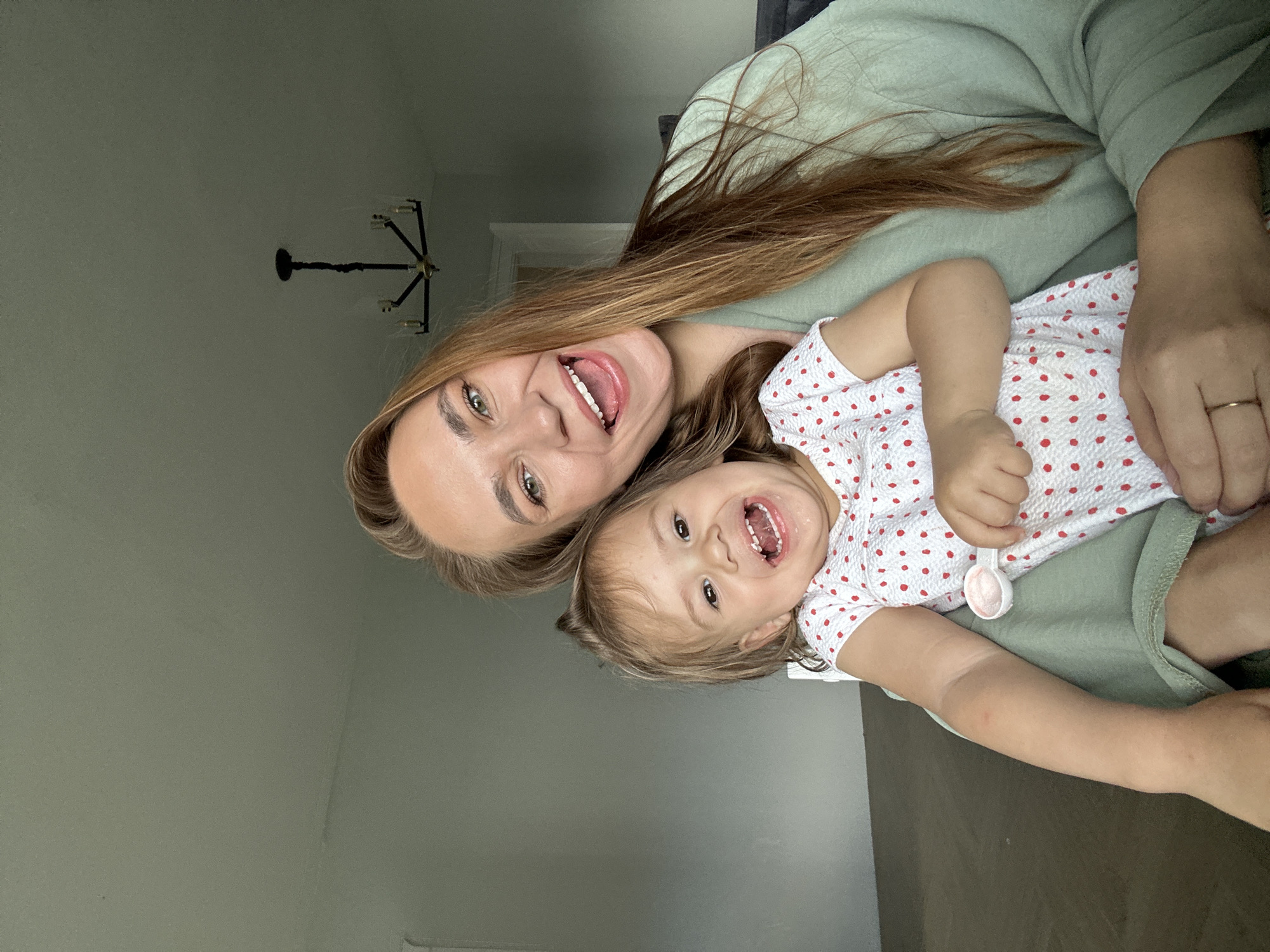 A woman and a young child are seated, with the child on the woman's lap, captured in a selfie-style photograph. The woman, with long, auburn hair, is looking at the camera with a wide smile. She is wearing a light green robe. The child, facing the camera, also has a broad, toothy smile and is wearing a white romper with red polka dots. The child is holding a small white spoon. They are indoors, with a plain gray wall behind them and a chandelier visible on the ceiling. The floor is covered in what appears to be dark wood flooring. The scene is likely a residential setting. There is no information to suggest this location is in Kharnavy, Russia.