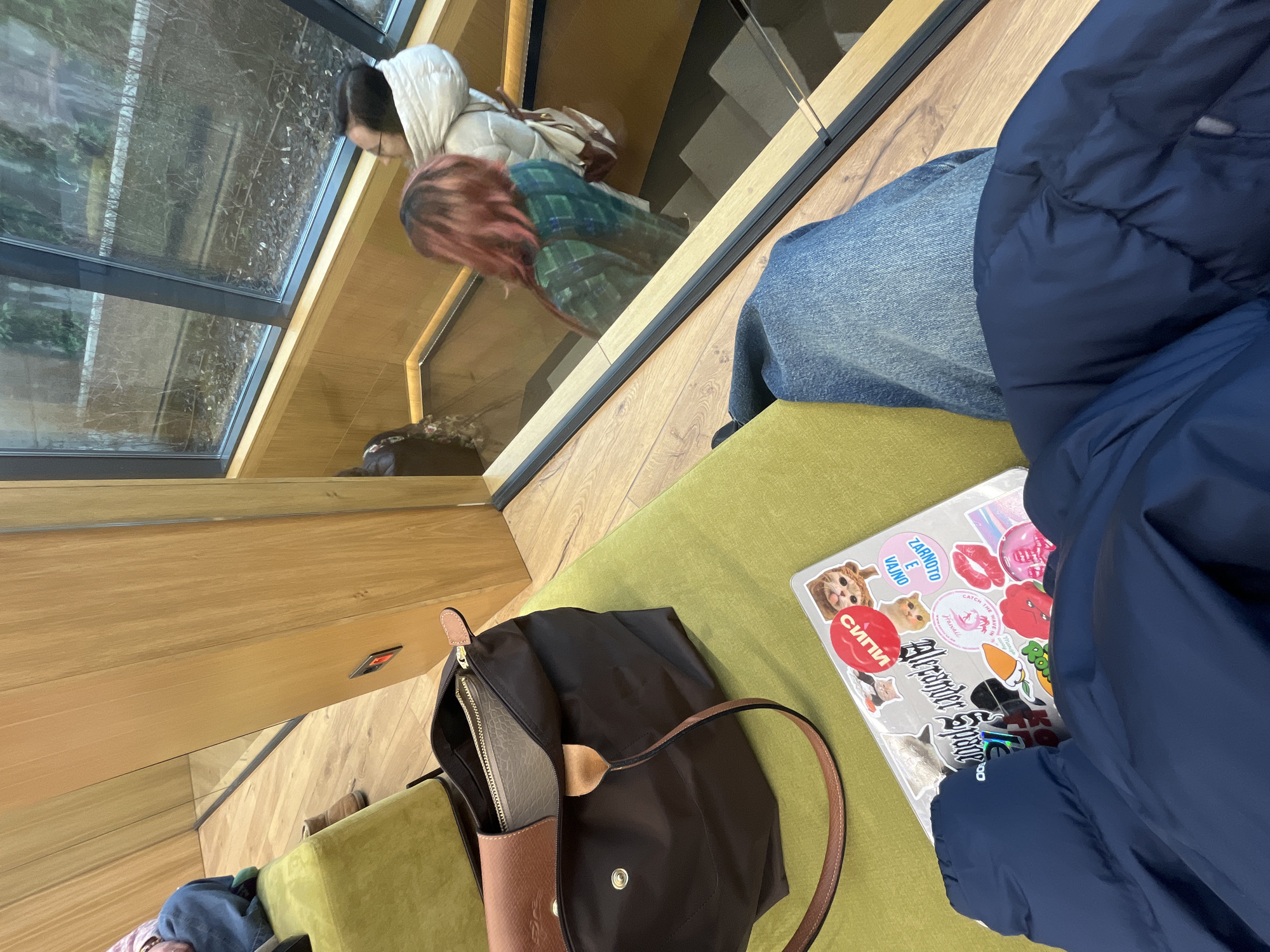 The image shows an interior scene. A person wearing jeans and a large, dark blue puffer jacket sits on a green cushioned bench. On the bench next to the person is a laptop covered in stickers, and a dark brown Longchamp tote bag is also placed next to the laptop. A window provides a reflection of two people outside. The reflection shows a woman with red hair wearing a green plaid jacket and another woman wearing a white hooded jacket. The location is likely in Sofia, Bulgaria as indicated by the Cyrillic script on one of the stickers on the laptop. The interior space seems modern with wooden accents.