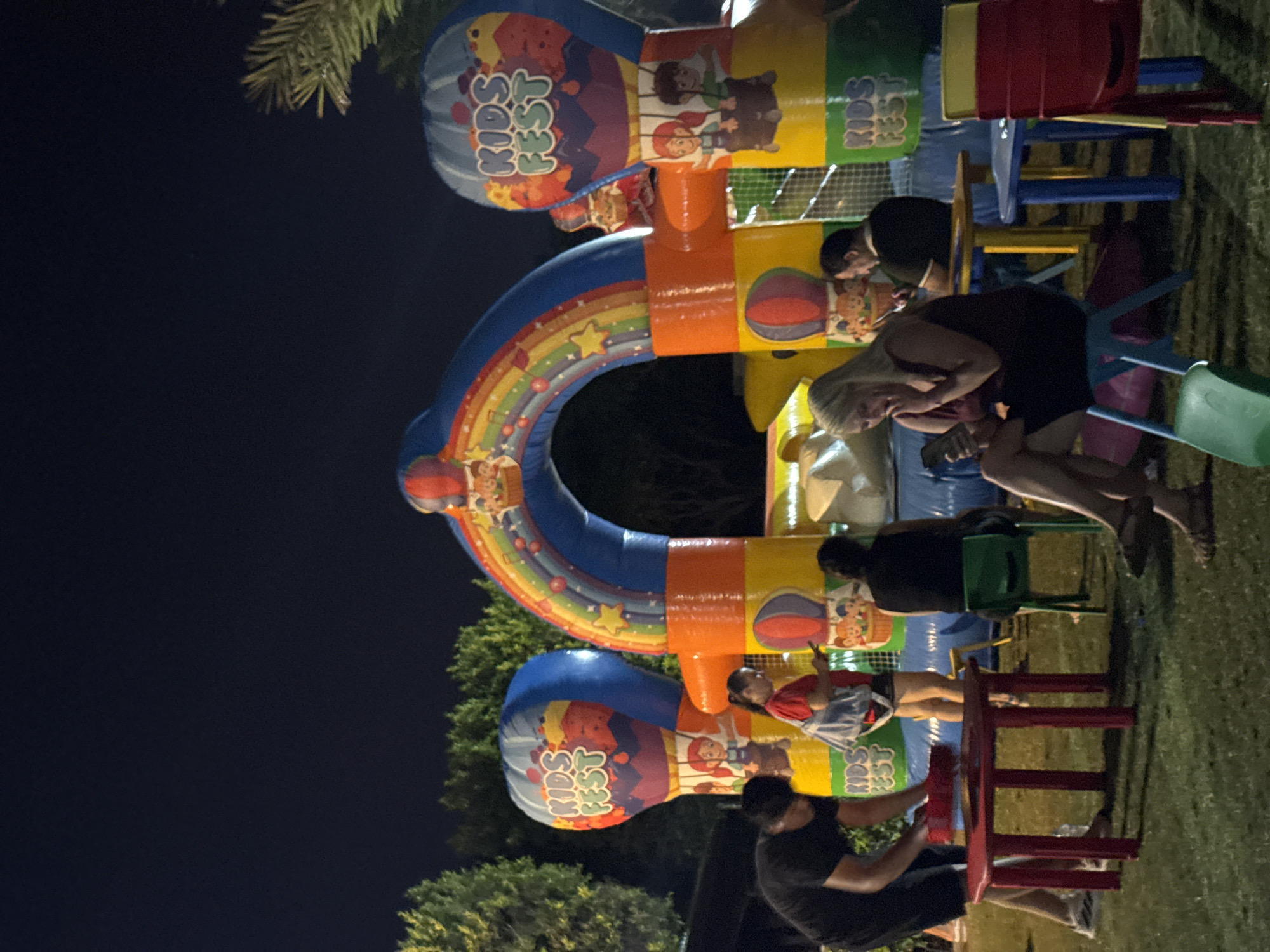 In Aksu, Turkey, at nighttime, an inflatable kids' festival attraction featuring hot air balloons and a rainbow arch is illuminated. Several people are present in front of the attraction. A man in a black t-shirt and shorts is standing by a red table, appearing to be setting something down. A woman in a red shirt and shorts is standing nearby, looking at her phone. Another woman, wearing a dark dress, is seated on a blue chair, also looking at her phone. Behind her, a man in a black shirt is seated at a yellow table. In the foreground, a green chair and several colorful chairs are visible, along with parts of tables. The ground is covered in grass, and trees are visible in the background.
