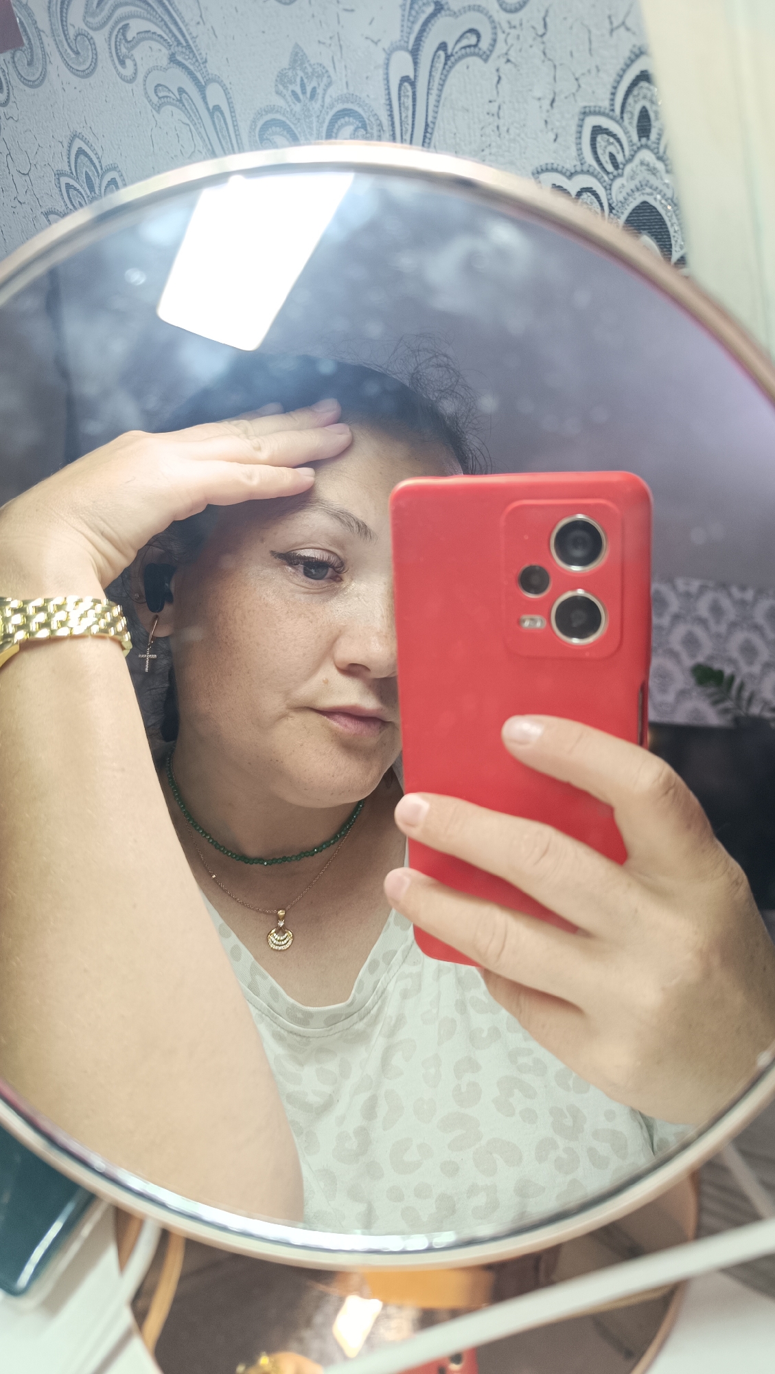 A woman is taking a mirror selfie in a bathroom in Chaikovsky, Russia. She is looking at the camera and has her left hand on her forehead. She is wearing a white t-shirt with a leopard print, a green beaded necklace, a gold bracelet, and earrings. The phone she is using is red. The background is a patterned wallpaper and a light fixture is reflected in the mirror.