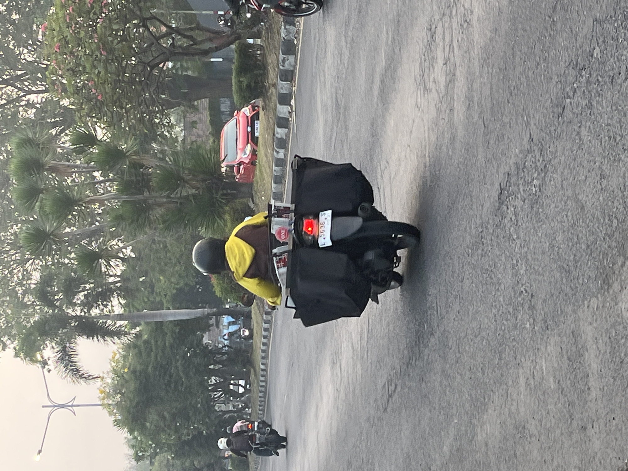 A rear view of a person on a motorcycle is captured from behind in Kludan, Indonesia. The person is wearing a yellow hooded jacket and a black helmet. The motorcycle has a large black cargo box mounted on the rear. In the background, other motorcycles are visible, along with trees, a red car, and a streetlamp. The road appears to be asphalt, and there is a curb on the right side of the image. The lighting suggests it may be early morning or late afternoon.