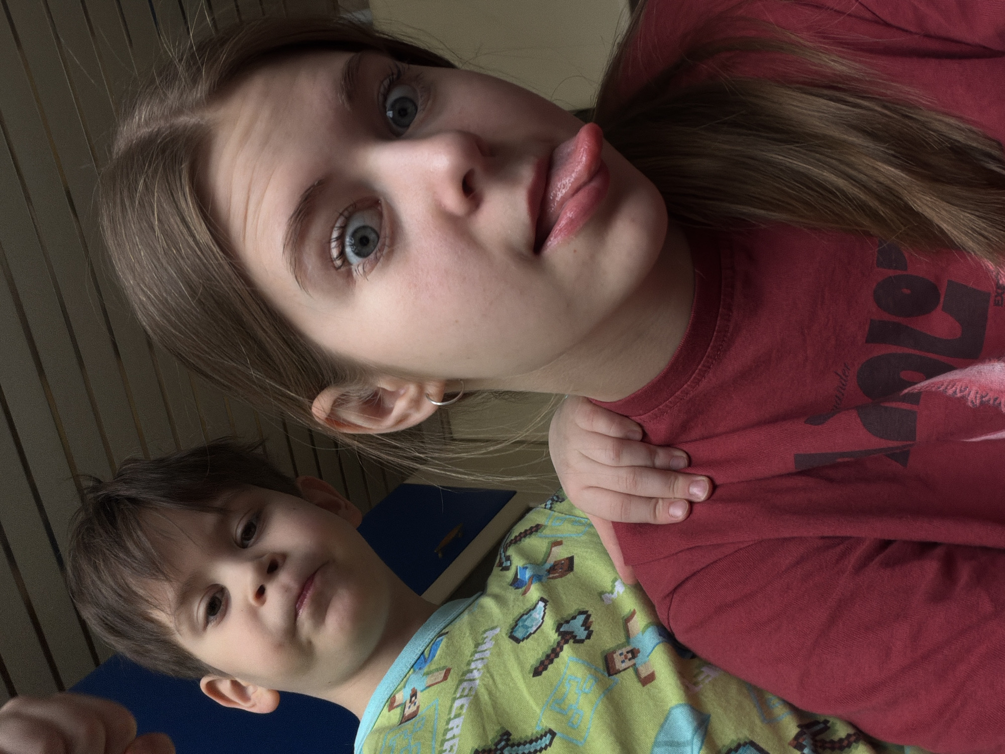 Two individuals are depicted in an indoor setting. The foreground shows an adolescent female, possibly early to mid-teens, with fair skin, light brown hair, and light-colored eyes. She is sticking out her tongue and looking at the camera. She wears a red t-shirt. Adjacent to her is a younger boy, approximately six to eight years old. He has brown hair and is wearing a green shirt with Minecraft-themed patterns. The location may be a private residence, potentially in Perm, Russia, though there is no concrete evidence to confirm this. The background shows a wall. Other objects include a blue surface. There are no clear actions or interactions beyond posing for the photograph.