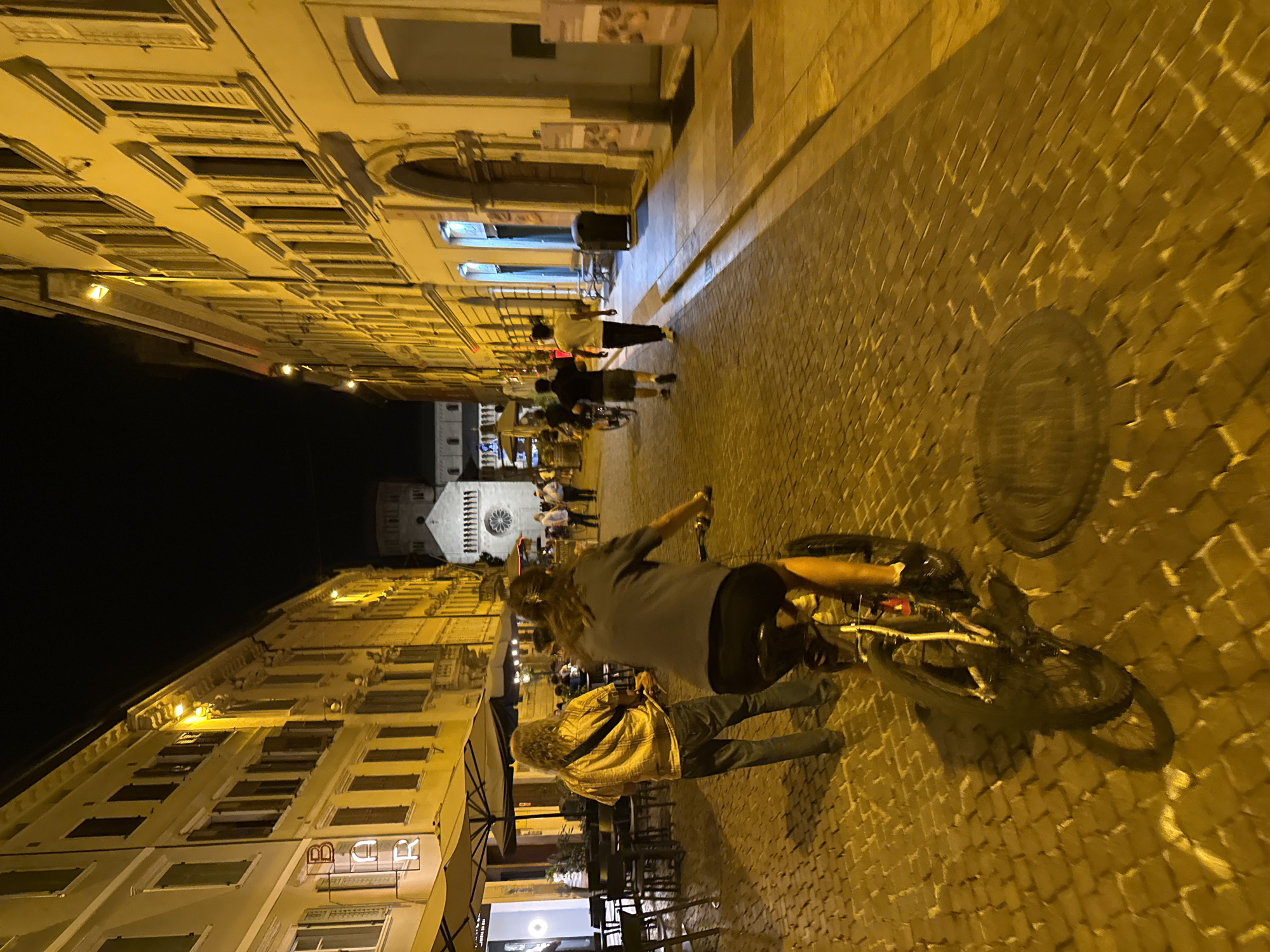 The image depicts a street scene at night in the city of Trento, Italy. The street is paved with cobblestones and illuminated by warm, yellow streetlights, casting long shadows. On the left, buildings with arched windows and shuttered openings line the street, with a neon "BAR" sign visible. On the right, similar buildings also feature arched doorways and windows.
In the foreground, a person is riding a bicycle, their body angled away from the camera. Their legs are visible, and they appear to be wearing dark clothing. The bicycle's wheels and frame are detailed, with shadows cast by the bike onto the cobblestones. Behind the cyclist, a person with long, curly hair wearing a patterned shirt and jeans stands facing away from the camera, holding a stick or a drumstick. Further down the street, several other individuals are walking or standing, some in groups, and at least one other person appears to be on a bicycle in the middle ground. In the background, a large, historic building with a circular window and a tower is visible, likely a church or cathedral. The overall atmosphere is that of a lively, pedestrianized area in an Italian city at night.