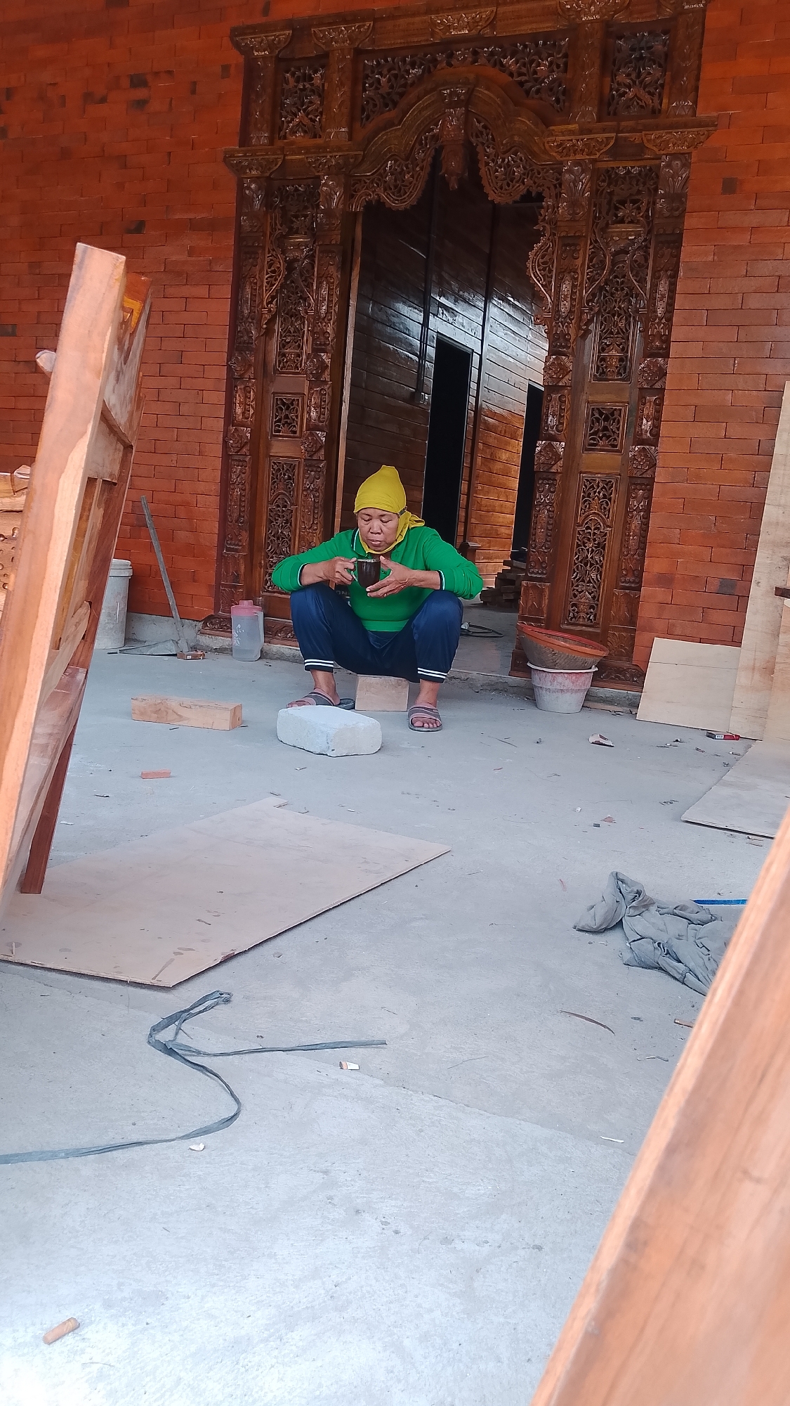A person wearing a yellow headscarf and a green sweatshirt is squatting on a piece of wood and drinking from a cup in Karanglangu, Indonesia. The person is in front of an elaborately carved wooden doorway with a brick wall to the left. Plywood sheets are on the ground in front of the person, along with a white rectangular object and a piece of lumber. To the right of the person are some buckets and fabric. The ground is concrete.