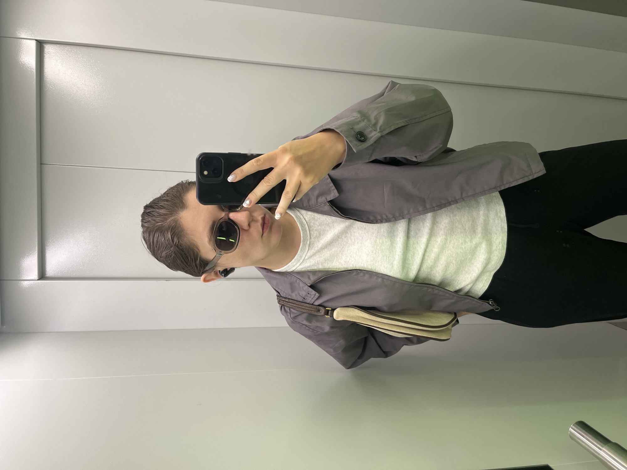 A person stands in what appears to be an elevator with white panel walls. The person is taking a selfie with a black smartphone. They wear sunglasses, a white t-shirt, and a gray jacket, with black pants. They have an earbud in their ear and a purse or bag with a beige strap is visible. The location is unknown, and there is no information to suggest it is in Krasnoyarsk, Russia.