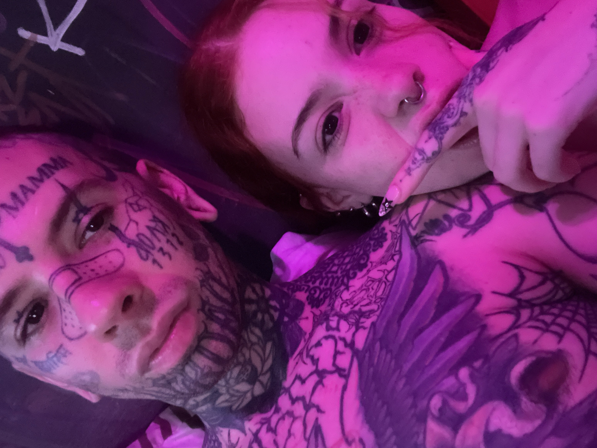 The image shows two individuals lying down, close to each other. The person on the left has extensive tattoos on their face, including the word "MAMMA" on their forehead and other symbols/numbers. The individual on the right has red hair and is wearing a nose ring and earrings. They have tattoos on their hand and chest. The scene is lit with a pinkish light. No location information is discernible.