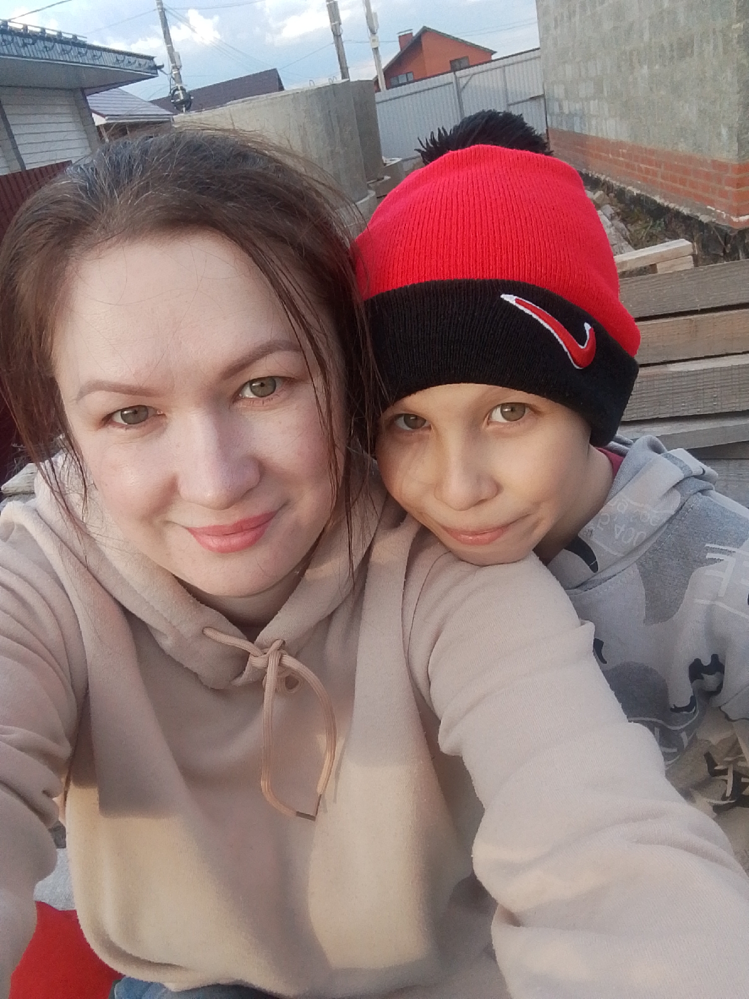A selfie shows a woman and a child outdoors in Прикамский, Russia. The woman, on the left, wears a beige hoodie and has brown hair. The child, on the right, wears a grey camouflage hoodie and a red and black Nike beanie. They are standing close together, and both are looking at the camera with slight smiles. The background shows a brick wall and other buildings, suggesting a residential area. The lighting indicates it might be late afternoon.