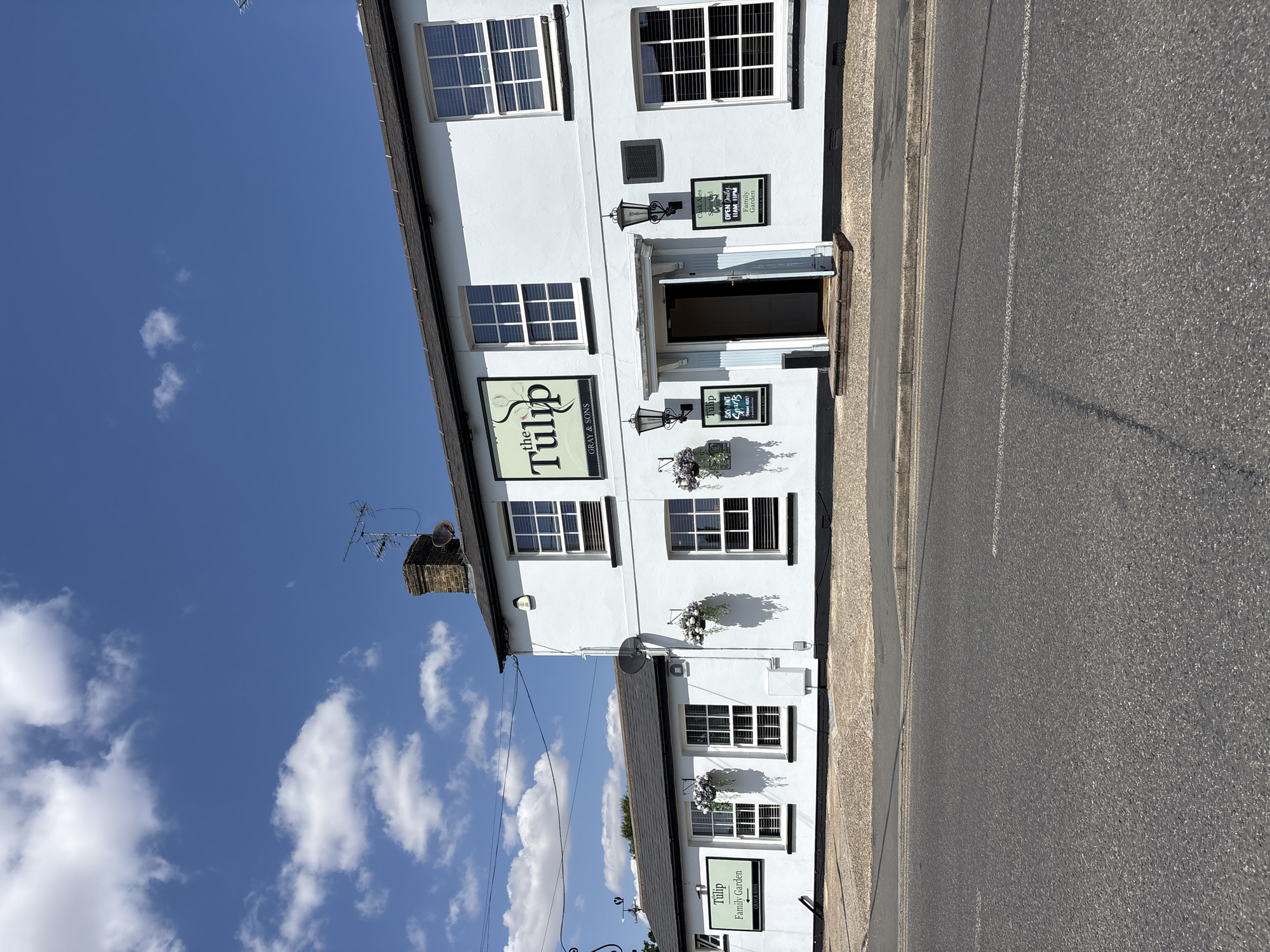A white two-story building with a dark roof and a brick chimney is pictured on a sunny day. The building has several windows with white frames and blinds. Signs on the building indicate it is "the Tulip" with "GRAY & SONS" written below. One sign also advertises "Family Garden" and "Craft Ales". The building is situated on a street with a tarmac road in the foreground. There are hanging flower baskets on the exterior of the building. The sky is blue with scattered white clouds.