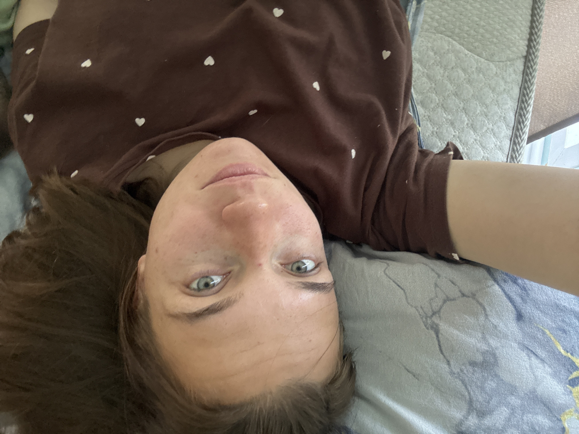 A person with fair skin, brown hair, and light-colored eyes is lying on a bed. The person is wearing a brown shirt with small white heart patterns. The bed has a light-colored patterned sheet and a portion of a light grey headboard is visible in the upper right corner. The location of the photo cannot be determined to be Раздолье, Russia from the visual information provided.