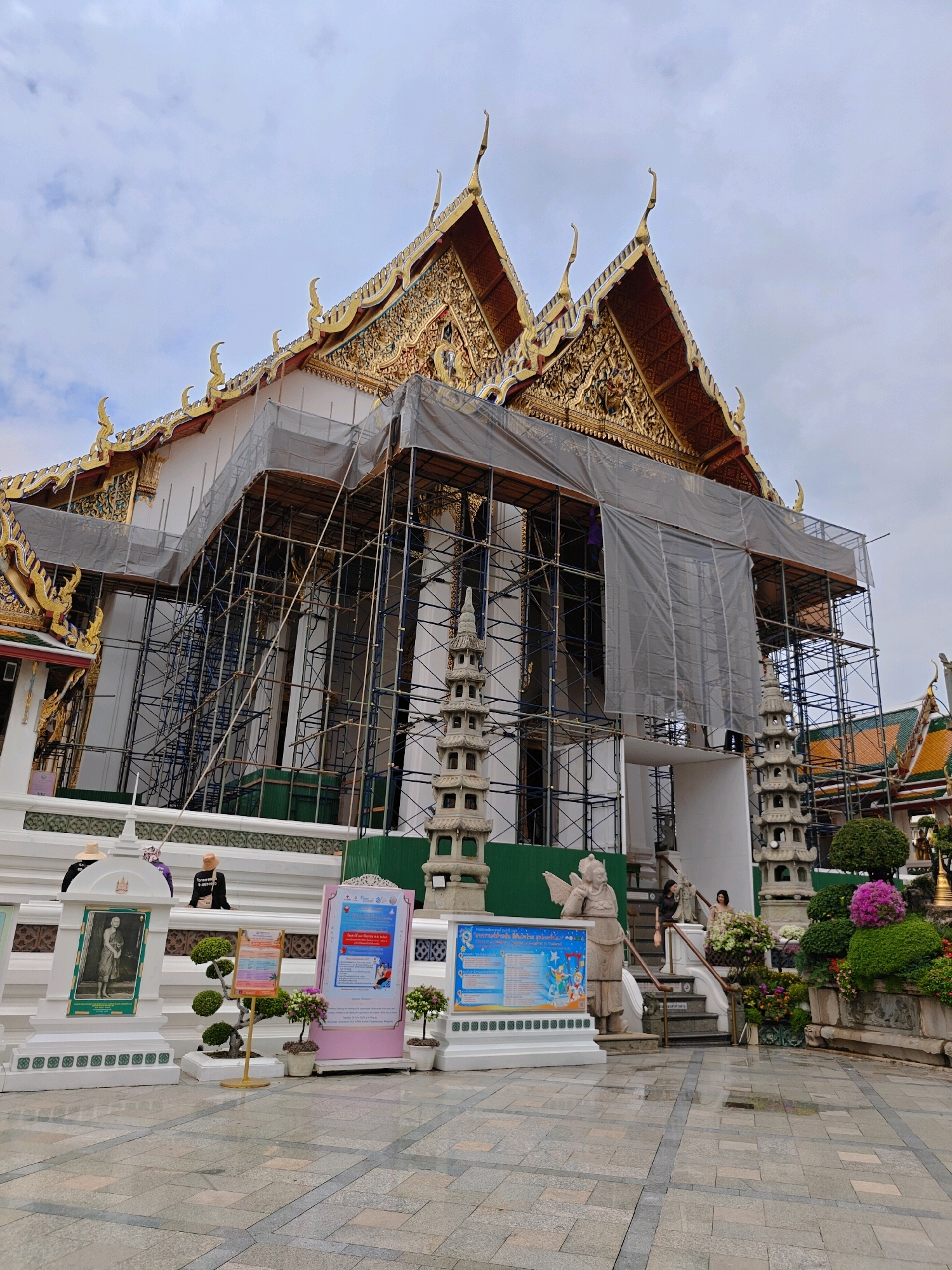 The image captures a side view of a temple in Bangkok, Thailand, which is undergoing renovations. The temple's ornate golden roof, adorned with intricate carvings and decorative elements, is visible against a cloudy sky. Scaffolding and tarpaulins cover a significant portion of the temple's exterior, indicating ongoing construction or repair work. Several people are present in the scene, some of whom are seated on steps, while others are walking or standing. Various decorative elements, including stone statues, potted plants, and informational displays, are placed around the temple grounds, suggesting a cultural or tourist site. The ground is paved with patterned tiles, and the overall atmosphere appears to be overcast.