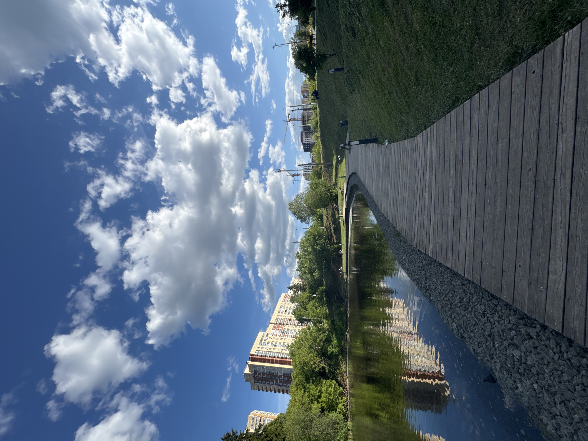 The image displays an urban park scene in Izhevsk, Russia, under a clear blue sky with scattered white cumulus clouds. The foreground features a dark wooden boardwalk curving along the edge of a calm body of water, likely a pond or river. Between the boardwalk and the water, a narrow strip of dark grey riprap or gabion stones stabilizes the bank. To the right of the boardwalk, a manicured grassy slope ascends, dotted with several dark, modern-style path lights and smaller green shrubs.
In the mid-ground, across the water, dense green deciduous trees line the opposite bank. Behind these trees and further to the left, several large multi-story residential apartment buildings are visible. These buildings exhibit light-colored facades with prominent reddish-brown or orange vertical and horizontal accents. Their reflections are visible on the water's surface. On the far side of the water, on a grassy area, several indistinct human figures are present, some appearing to be seated or standing near park amenities.
Towards the right mid-ground and background, beyond the initial grassy slope, more urban structures and several prominent construction cranes indicate ongoing development. The overall lighting suggests a bright daytime setting, likely during spring or summer, given the lush green foliage. No specific actions or interactions between individuals are clearly discernible due to distance.