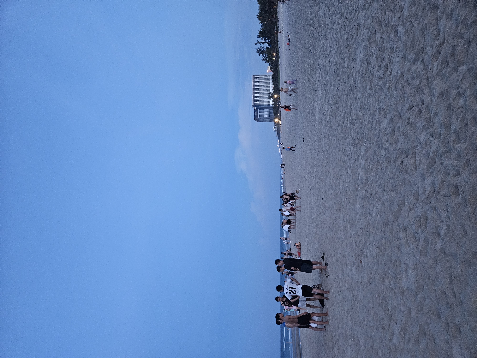 The image depicts a beach in Da Nang, Vietnam, likely My Khe Beach given the presence of large hotels in the background. It's dusk, and the sky is a deep blue with some faint clouds. The beach is sandy, with gentle waves lapping at the shore.
In the foreground, a group of young men are standing and talking. One of them is wearing a white jersey with the number "21" on the back. They are all dressed in casual beach attire, like shorts and t-shirts.
Further down the beach, there are other people scattered about. Some are walking along the shoreline, while others are playing or relaxing on the sand. In the distance, two large, modern hotels are visible, suggesting a developed tourist area. Palm trees line the edge of the beach, adding to the tropical atmosphere. The overall impression is one of a relaxed evening gathering on a popular beach.