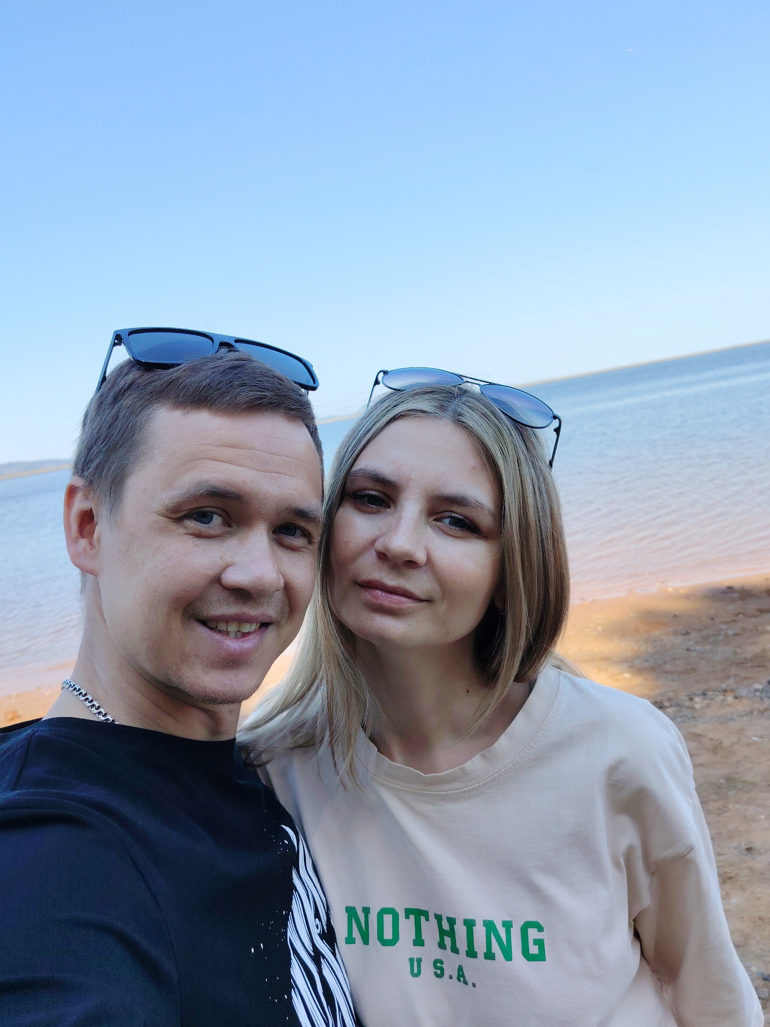 A man and a woman are taking a selfie in front of a body of water under a clear, blue sky. The man is on the left, wearing a black t-shirt, and has sunglasses perched on his head. He has short hair and is smiling. The woman is on the right, wearing a light beige t-shirt with the word "NOTHING" printed in green along with the letters "U.S.A." underneath. She also has sunglasses on her head and is looking at the camera. The background includes a sandy or gravelly area near the water's edge. The location is Chaikovsky, Russia.