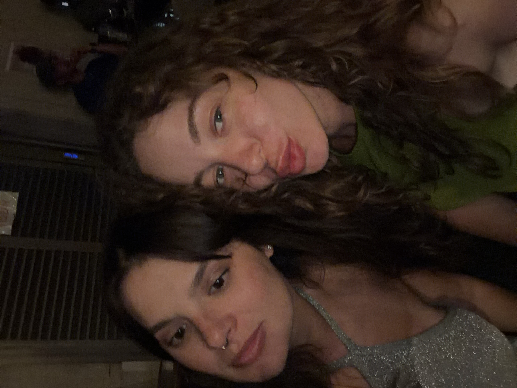 The image shows a close-up selfie of two young women in a dimly lit indoor setting. The woman on the left has dark hair, a nose ring, and is wearing a sparkly silver top. The woman on the right has curly brown hair and is wearing a green top, puckering her lips. In the background, a person in a blue shirt is visible, along with some out-of-focus objects that suggest a bar or restaurant environment. The lighting is warm and appears to be artificial, creating shadows and highlighting the subjects' faces. There are no specific visual cues within the image to determine a location in Tel-Aviv, Israel.