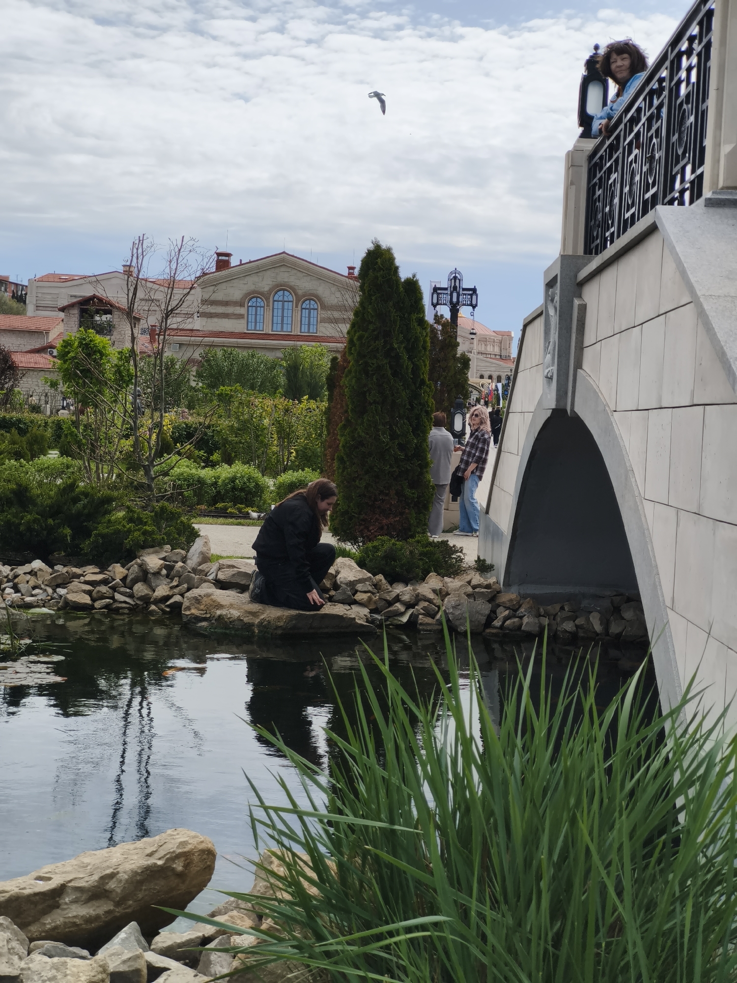 The image captures a serene outdoor scene in what appears to be a park or garden setting. A woman is crouched on rocks near a pond, with her gaze directed towards the water. Her reflection is visible in the pond's surface. The pond is surrounded by rocks, greenery, and tall green plants in the foreground. In the background, there is a bridge with people visible on it, a building with arched windows, trees, and other landscaping elements. The sky is overcast. The location is in the city of Sevastopol, Ukraine.