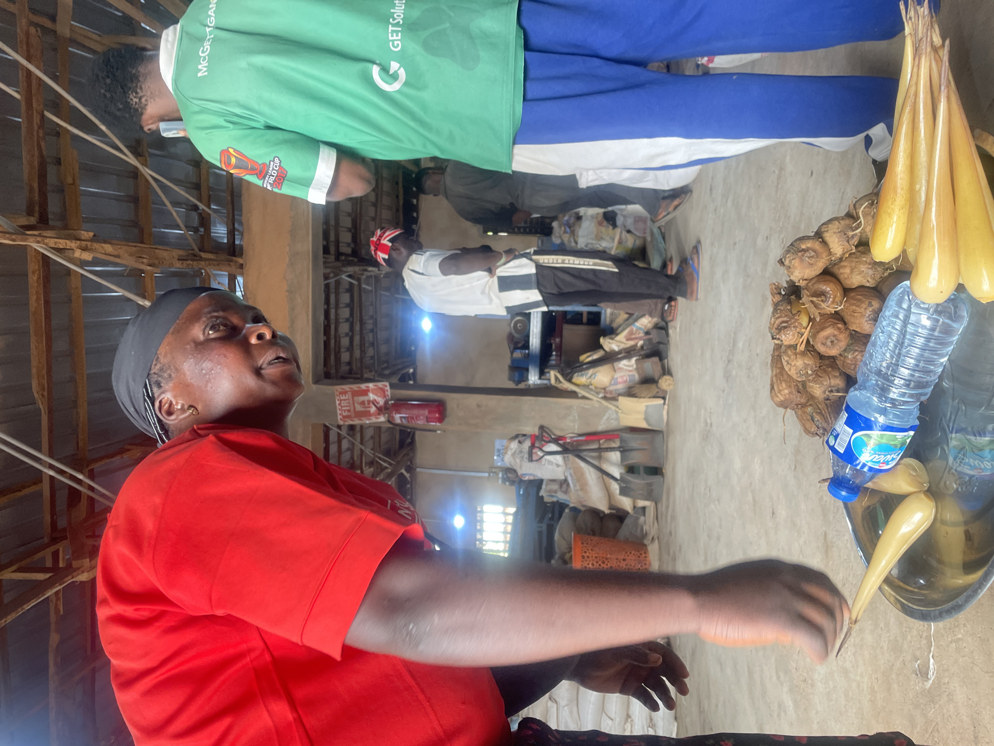 The image depicts a scene inside a market or warehouse in Gura Top, Nigeria. In the foreground, a person wearing a red shirt is leaning over a silver tray containing yellow, elongated root vegetables, a bottle of water, and a pile of brown, dried root vegetables. The person's face is partially visible, showing them looking downwards. To the right, another individual is visible from the back, wearing a green shirt with "McGETTGANDA" and "WORLD CUP 2017" printed on it, along with blue pants. In the background, several other people are present, along with sacks, tools like shovels and a broom, and a fire extinguisher mounted on a wall. The structure appears to be made of wood and corrugated metal, suggesting an open-air or partially enclosed market setting. The overall lighting is dim with some areas illuminated by artificial lights.