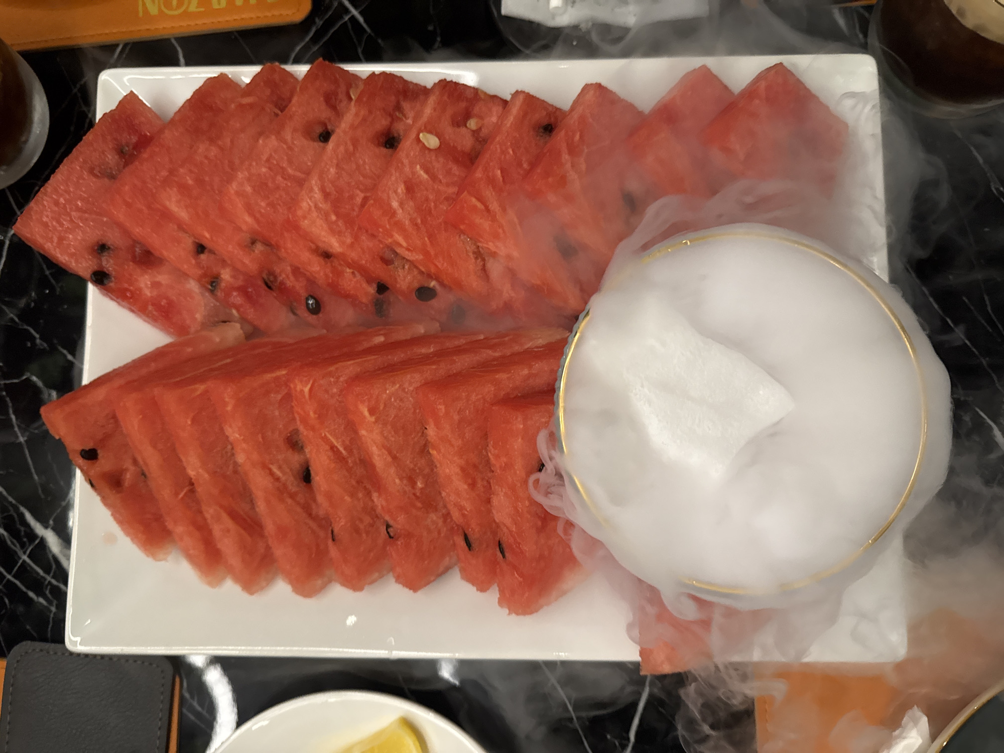 A top-down shot shows a rectangular white plate filled with watermelon slices, arranged in two rows, sitting on a black marble-like surface. White smoke, possibly from dry ice, is emanating from a round gold-rimmed bowl placed amidst the watermelon slices, partially obscuring the fruit. Some additional objects such as a menu, glasses and other plates are partially visible on the sides of the photograph. It is impossible to determine the location of the image within Yerevan, Armenia, based solely on the details provided.