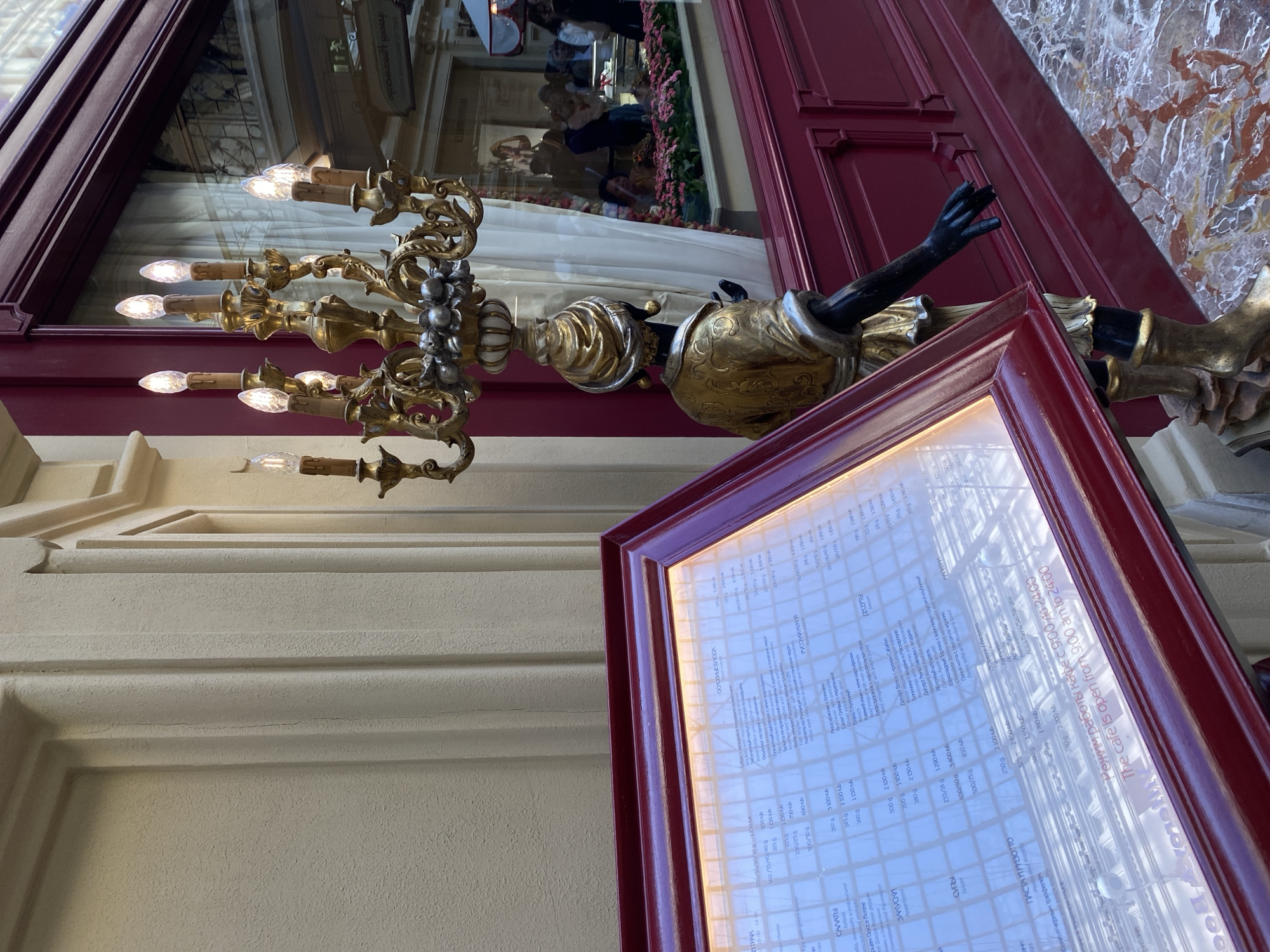 Here is a detailed description of the image:
**Scene:** An outdoor view of a restaurant's facade, likely in Moscow, Russia, based on the Russian language menu.
**Subjects:**
* An ornate candelabra with black arms holding it.
* A framed menu displaying a variety of dishes and prices in Russian rubles, which suggests Moscow as the location.
* People are reflected in the glass of a cabinet.
**Objects:**
* Menu board with a burgundy frame, displaying the restaurant's offerings.
* The candelabra features a gold and silver colored design, with lit bulbs at the ends of each branch.
* The restaurant's exterior wall, painted in off-white color.
* A red colored door/wall.
**Environment:**
The image appears to be taken during the day, with natural light illuminating the menu board and the surroundings. The architecture has classic elements, suggesting a sophisticated and possibly historic district within Moscow.