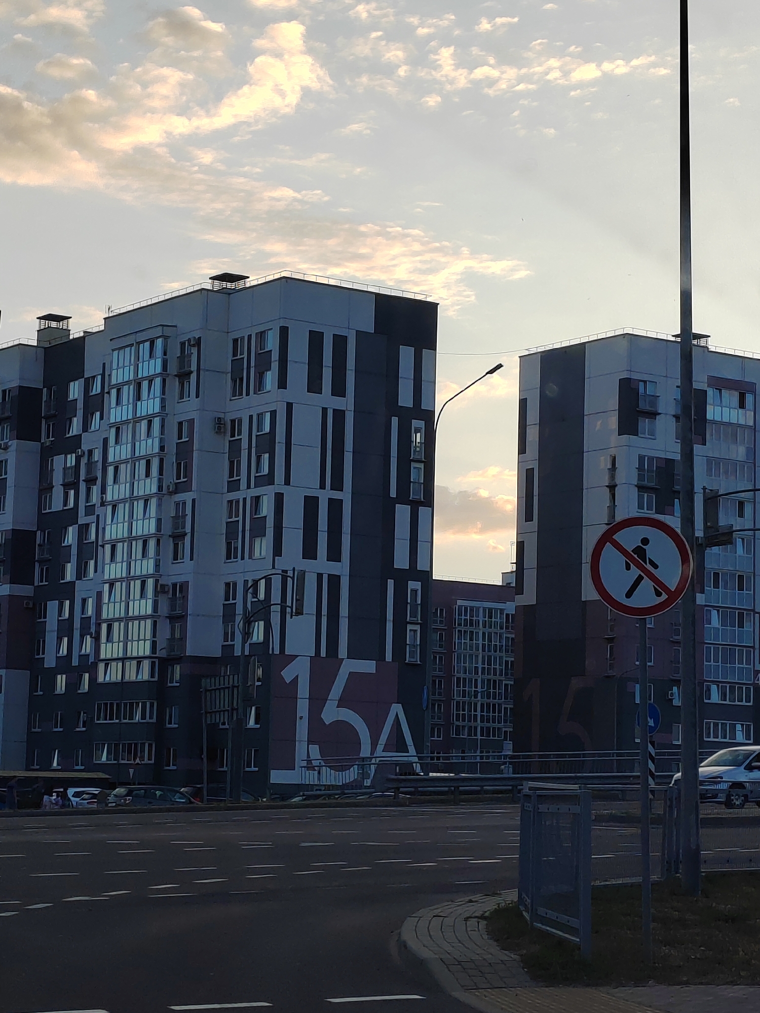 The image depicts a street scene in the city of Brest, Belarus. In the foreground, there is a road with white dashed lines marking lanes. To the right, a metal fence encloses a patch of grass and a sidewalk made of paving stones. Beyond the road, a pedestrian crossing sign with a red circle and a diagonal line over a silhouetted figure is visible, indicating no pedestrians.
Dominating the background are two modern apartment buildings. The building on the left is darker with lighter grey and white vertical stripes, and features numerous windows. The building on the right is lighter in color with dark grey vertical accents and also has many windows. Large numbers "15" and "4" are painted on the side of the buildings. The sky above is partially cloudy, with sunlight filtering through, casting rays. A street lamp is visible to the left of the right building. Cars are parked or driving on the street in front of the buildings.
