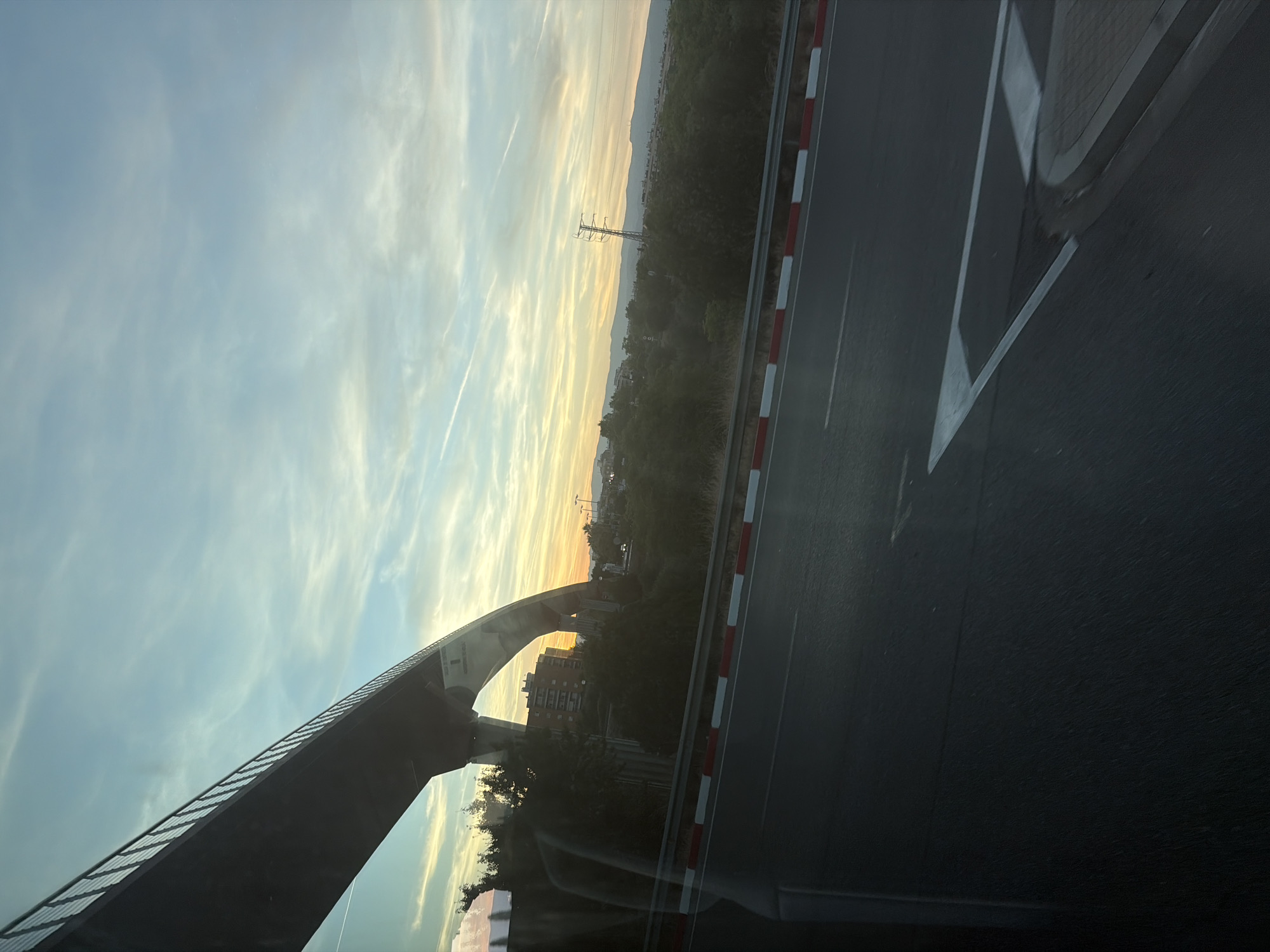 The image captures a road adjacent to a bridge structure with a natural landscape and sky in the background. The time of day appears to be sunset or sunrise, given the warm, golden hues in the sky and the soft lighting. The sky is a blend of light blue and yellow, with wispy clouds.
In the foreground, a dark asphalt road is visible, marked with white lines and bordered by a red and white striped barrier. The left side of the image is dominated by the underside of a large, modern bridge structure, which is dark and appears to be made of concrete or steel. The bridge arches over a natural area that includes trees and some buildings. A tall electrical pylon can be seen in the middle distance, silhouetted against the colorful sky.
The landscape beyond the immediate road and bridge features rolling hills or mountains in the far distance. There are hints of urban development with buildings visible amongst the trees. The overall mood is serene and perhaps a little melancholic, consistent with a twilight setting.
No people are visible in the image. The visible text appears to be a white label or sign on the underside of the bridge, but the text itself is illegible. The setting, with the bridge, road, and distant landscape, suggests an area on the outskirts of a town or city, likely in a developed region given the infrastructure. The weather appears to be clear, with the exception of the clouds. The location context provided is Vila-seca, Spain, which is consistent with the type of infrastructure and landscape depicted.