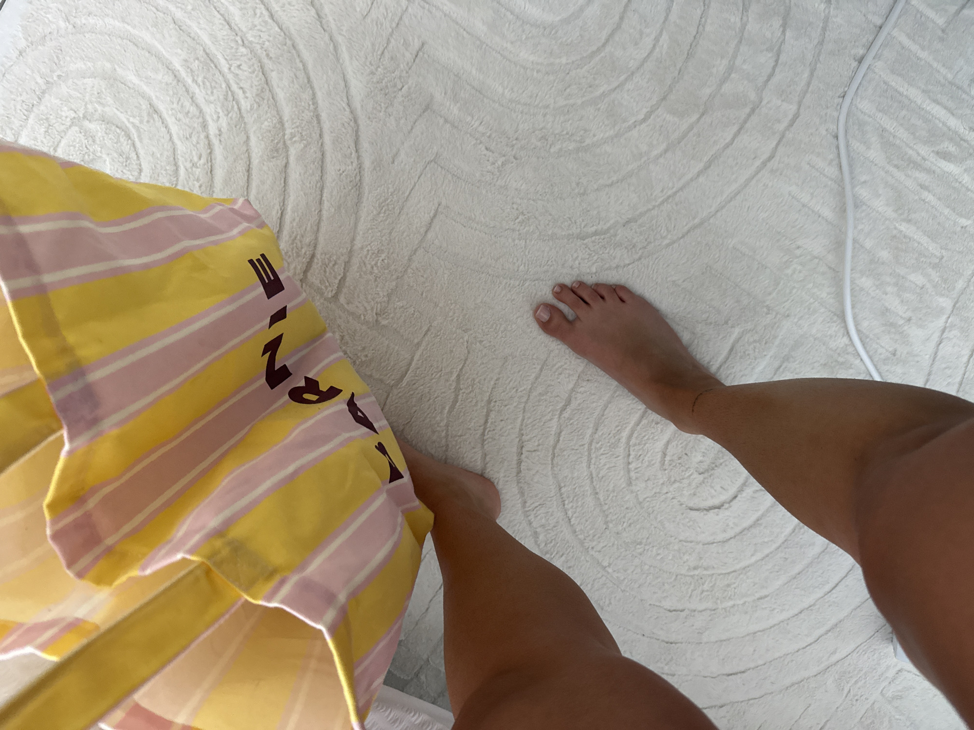 This overhead shot captures a person's bare, tanned legs and feet on a white, plush rug with a swirling textured pattern. The toenails are painted with a light polish. On the left, a yellow and light pink striped fabric item, likely a bag, is partially visible. Dark letters printed on this fabric clearly spell "ANIE", with hints of preceding letters that could form part of a word like "LANIE" or "FRANIE". A thin white cable or cord runs along the right side of the rug. The scene depicts an indoor setting, possibly a room in Cannes, France, hinting at a relaxed or vacation-like atmosphere consistent with the warm climate implied by the tanned skin. No specific activity is shown beyond the static pose.
