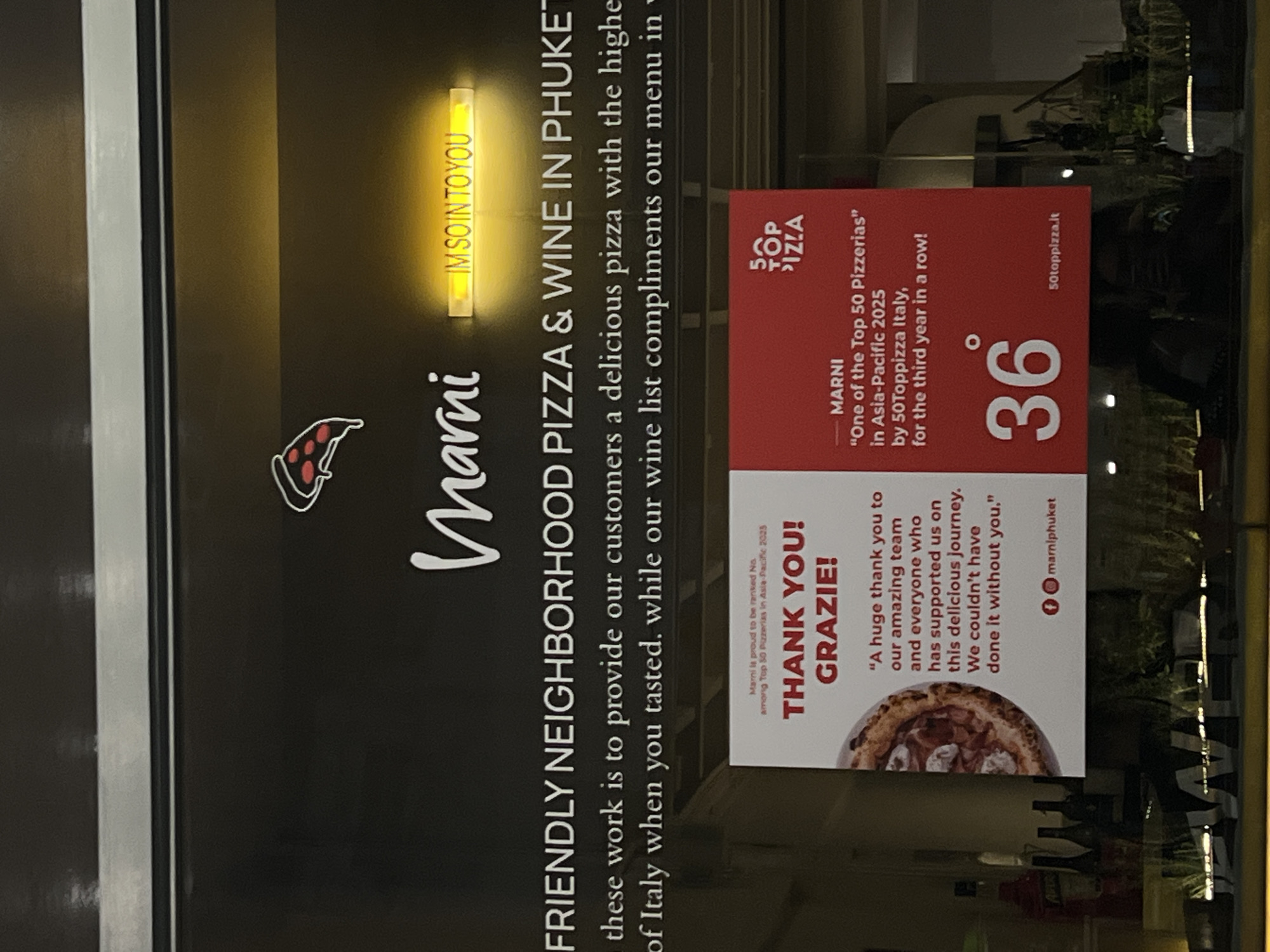 Here is a description of the image:
The image shows a restaurant window with a sign for "Marni Friendly Neighborhood Pizza & Wine in Phuket". The sign features a pizza slice graphic, the restaurant's name in cursive, and text describing their mission to provide delicious pizza and wine. A smaller sign highlights Marni's ranking among the Top 50 Pizzerias in Asia-Pacific for 2023 and 2025, with a "Thank You!" message and a picture of a pizza. The number 36 is displayed prominently on the sign, along with the website "50toppizza.it". The reflection in the window shows some of the interior of the restaurant and the street outside.