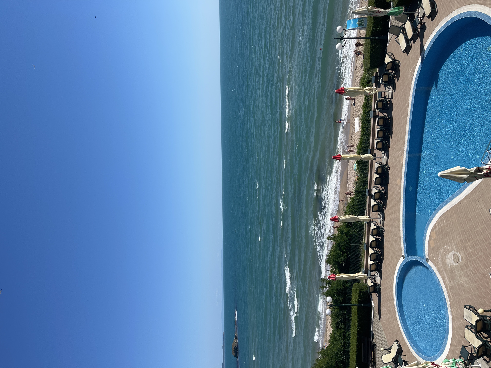 The image captures a sunny day at a coastal resort. In the foreground, a bright blue, free-form swimming pool is visible with lounge chairs surrounding it. Beyond the pool area, a sandy beach meets the turquoise ocean, where gentle waves are breaking. Several beach umbrellas are set up on the sand, and a few people are scattered along the shoreline and in the water. In the distance, the vast expanse of the sea stretches to the horizon under a clear blue sky, with a small rocky island visible on the left. The overall scene conveys a sense of relaxation and vacation.