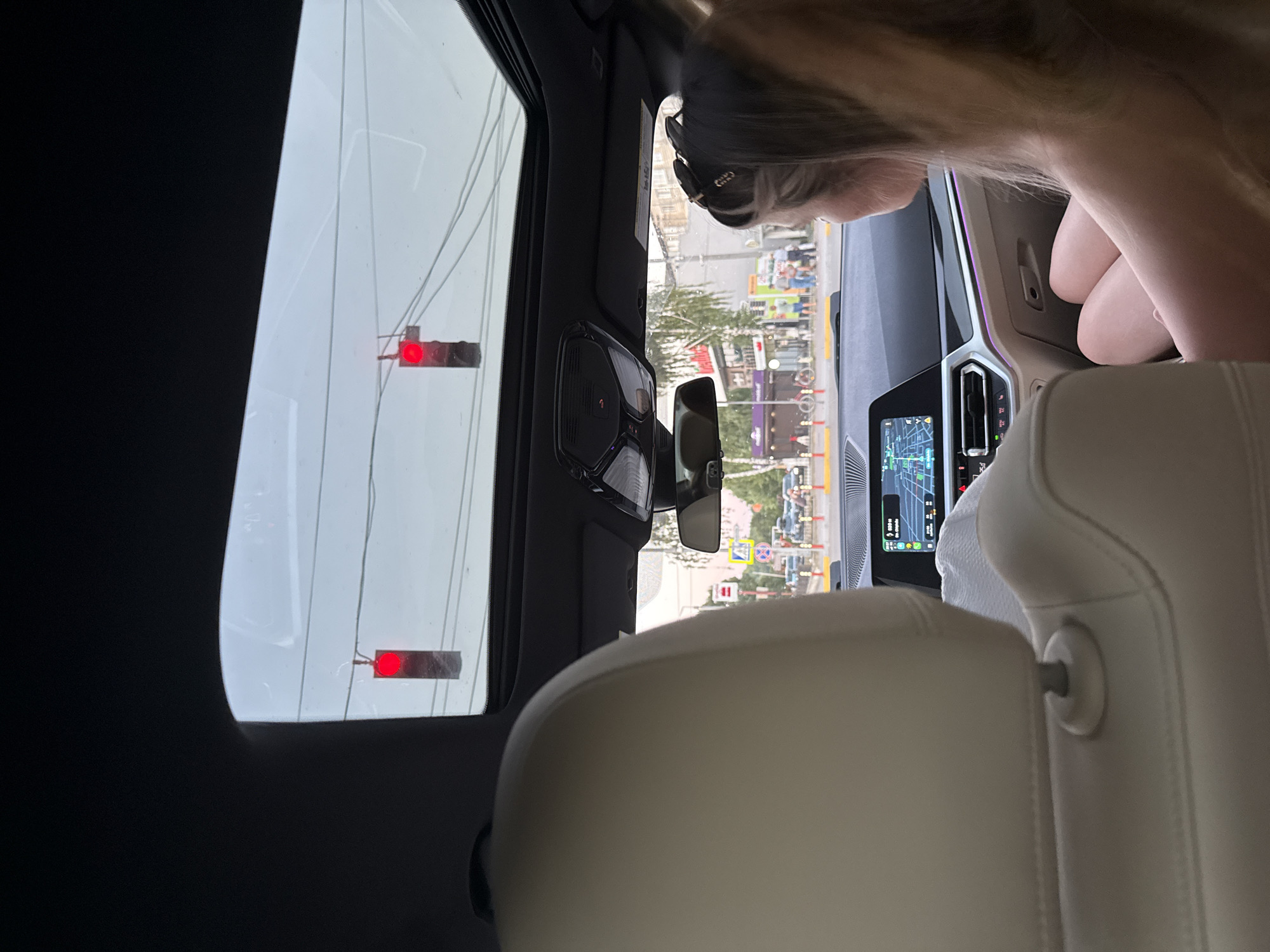 The image is taken from inside a car, looking out through the sunroof and windshield. The foreground shows the back of a car seat and a woman's head and shoulders as she looks out the side window. The middle ground shows a street scene with buildings, trees, and traffic. In the distance, there are more buildings and cars. The sky is overcast. There are two traffic lights visible through the sunroof, both showing red. The car appears to be stopped at an intersection. The woman in the foreground is wearing sunglasses on her head. The interior of the car is modern and luxurious, with a large infotainment screen displaying navigation. Based on the visible road signs and architecture, the location is identified as Fundul Galbenei, Moldova.