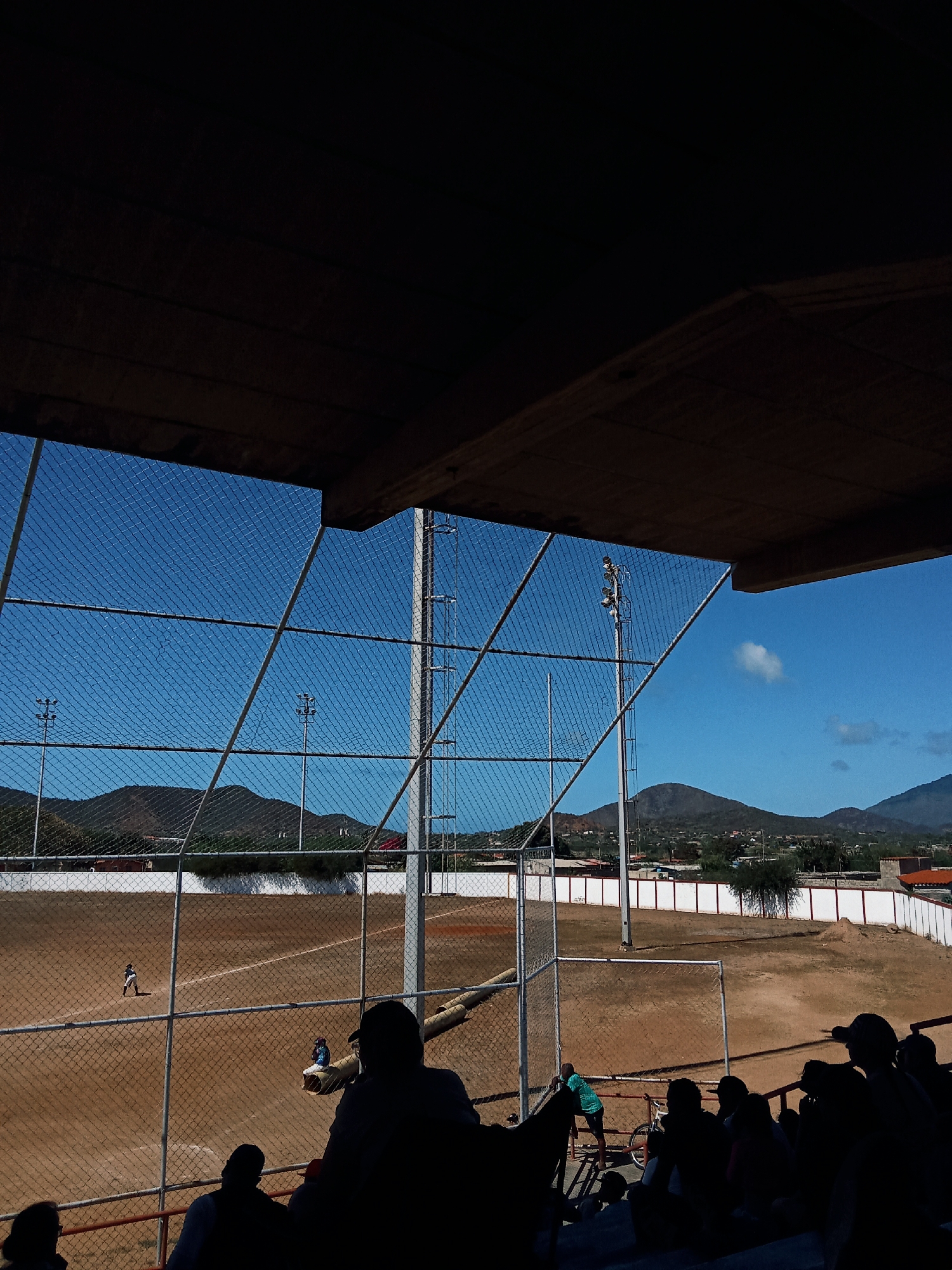 The image shows a baseball field in Juan Griego, Venezuela, viewed from under a roofed spectator area. A chain-link fence separates the viewers from the field. The playing surface is dirt, and a few people are on the field. A white wall surrounds the outfield. Utility poles with lights stand beyond the outfield fence. A few people are in the bleachers, watching. Behind the field, there are mountains and houses. The sky is clear and blue.