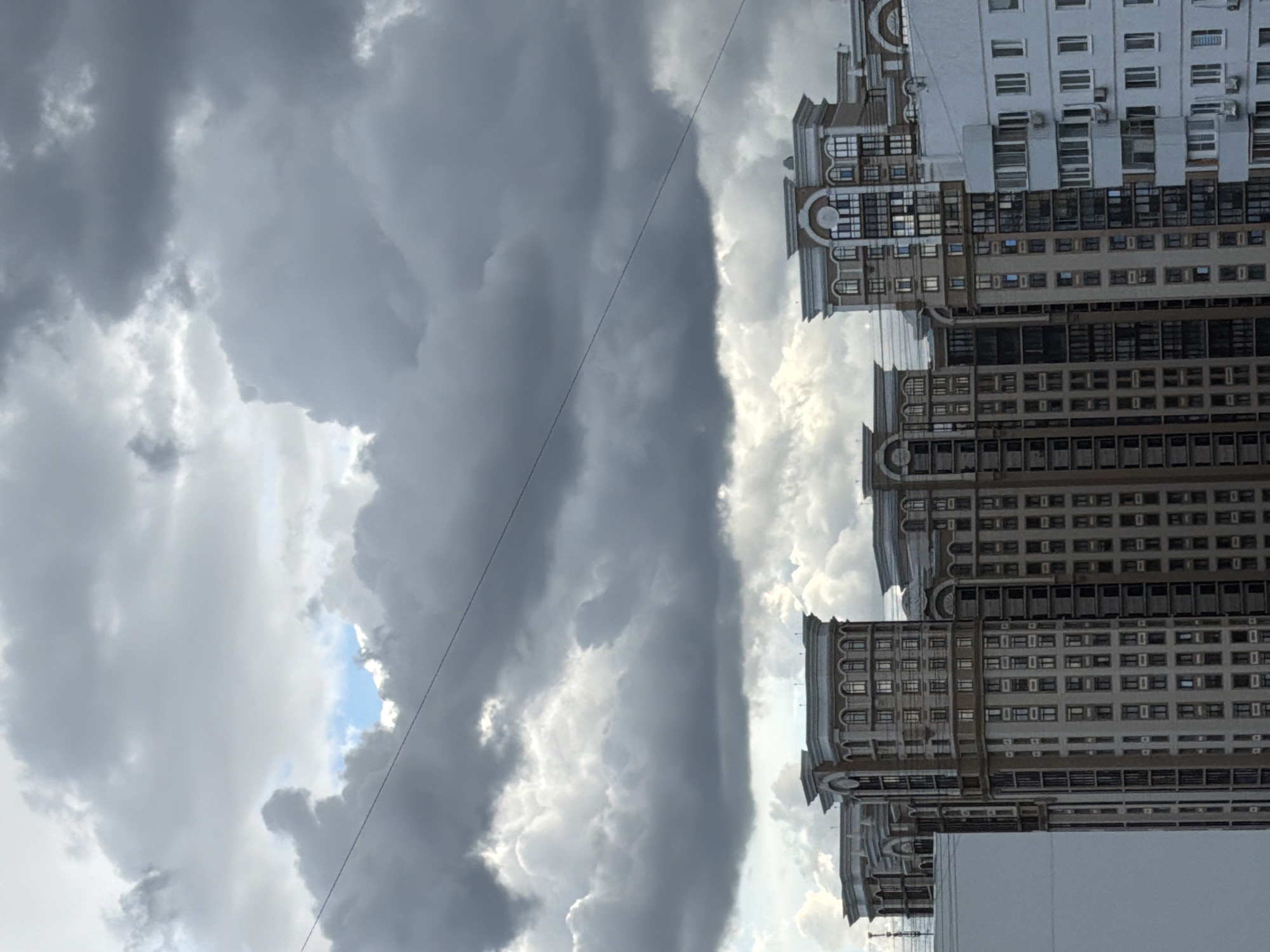 The image depicts tall apartment buildings against a stormy sky in Khimki, Russia. The buildings are multi-storied with repetitive window patterns and architectural details. The sky is filled with dark, billowing clouds, with a small patch of blue visible. A power line runs diagonally across the frame. The image is shot from a low angle, emphasizing the height of the buildings and the dramatic sky.