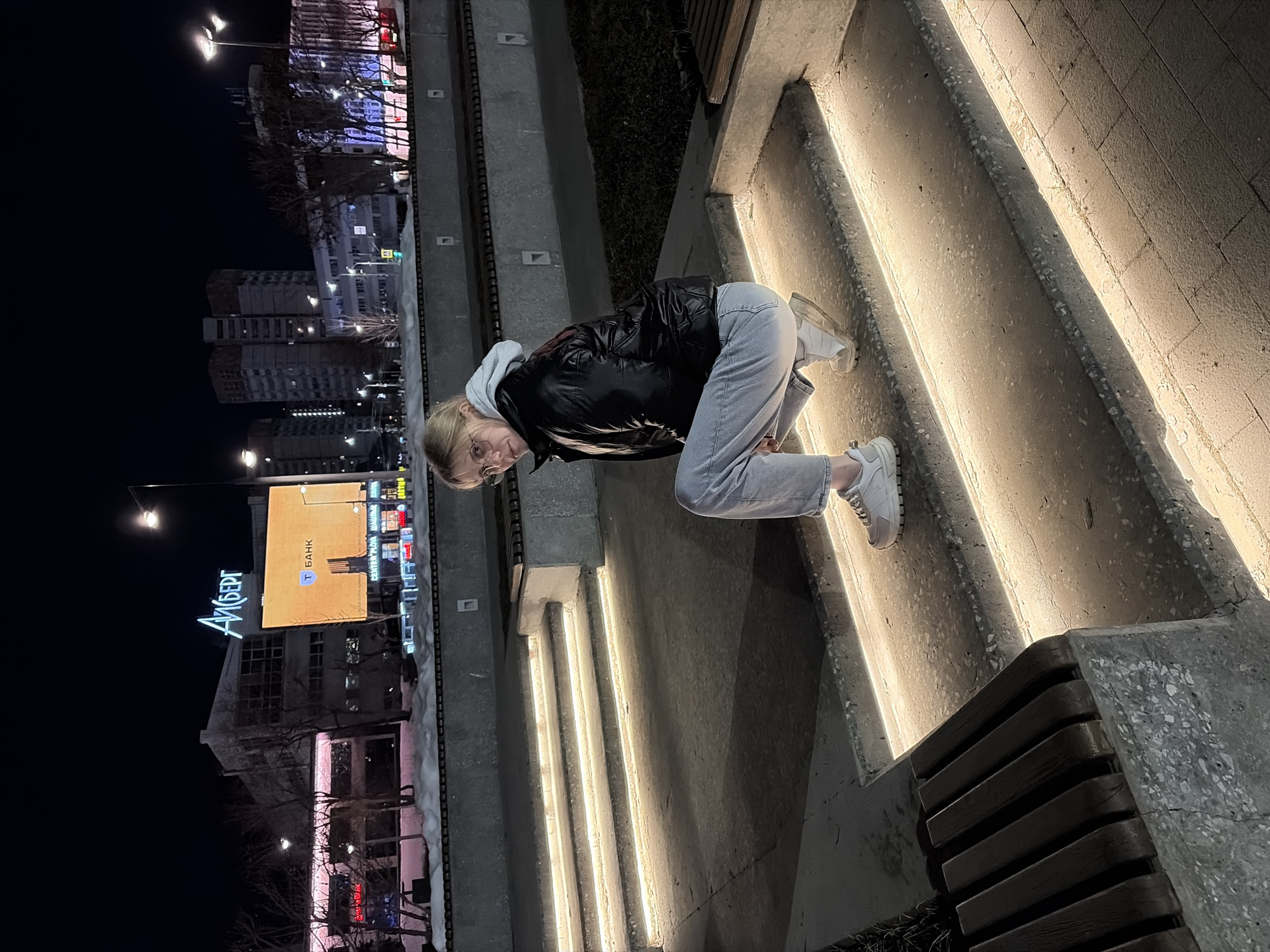 The image depicts a nighttime urban scene in Perm, Russia. A young adult female is the central subject, positioned in a squatting pose on illuminated concrete steps. She has blonde hair, wears glasses, a black puffer jacket, a light grey hooded sweatshirt, light blue jeans, and white sneakers. Her gaze is directed towards the camera.
The environment consists of a terraced public space featuring concrete steps, each illuminated along its edge by embedded linear LED lighting. A wooden slatted bench is visible to the left of the subject. Patches of snow are present on the ground, indicating a cold weather setting.
In the background, under a dark night sky, several multi-story buildings are illuminated. Prominently featured are the "АЙСБЕРГ" (ICEBERG) shopping center building, identified by its blue neon sign, and an adjacent large digital billboard displaying "Т БАНК" (T BANK) and "CENTER PLAZA." These landmarks pinpoint the location to a public area near the "Iceberg" shopping complex, likely around the intersection of ulitsa Lenina and ulitsa Popova or a nearby square in the city center of Perm. Streetlights and building lights contribute to the overall illumination of the urban backdrop. Bare trees are visible in the mid-ground.