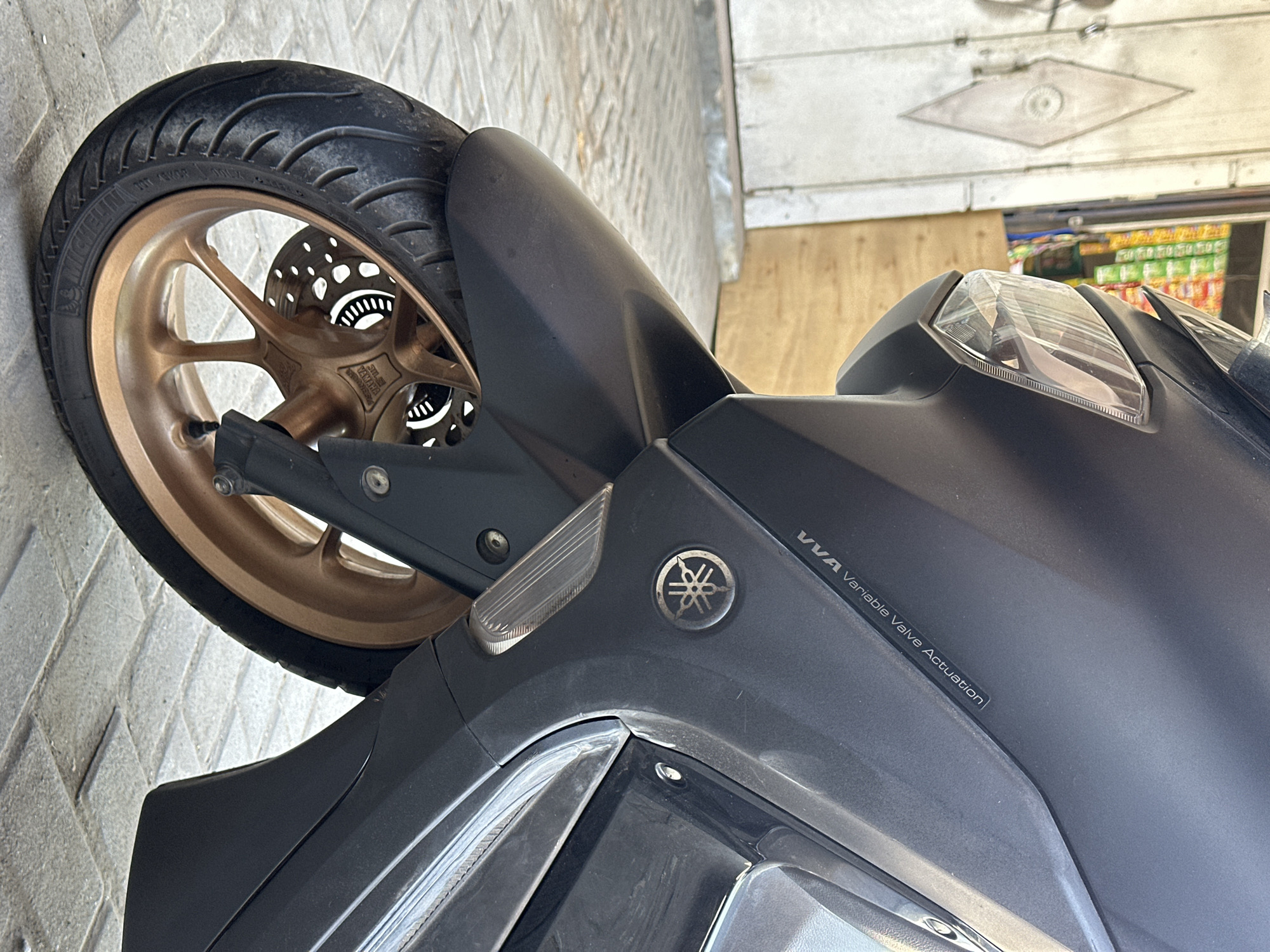 This is a close-up, eye-level, outdoor photograph of the front wheel and part of the fairing of a dark gray Yamaha NMAX motorcycle. The motorcycle is parked on a light gray, brick-patterned paved surface. In the background, to the left, is a white wooden door with a diamond-shaped decorative element. To the right of the door, a store display with rows of colorful products is visible. The motorcycle's front tire is a Michelin brand. The wheel rim is a metallic bronze color. The fairing displays the "VVA Variable Valve Actuation" text and a circular Yamaha logo. The scene is set in Bulakrejo, Indonesia.