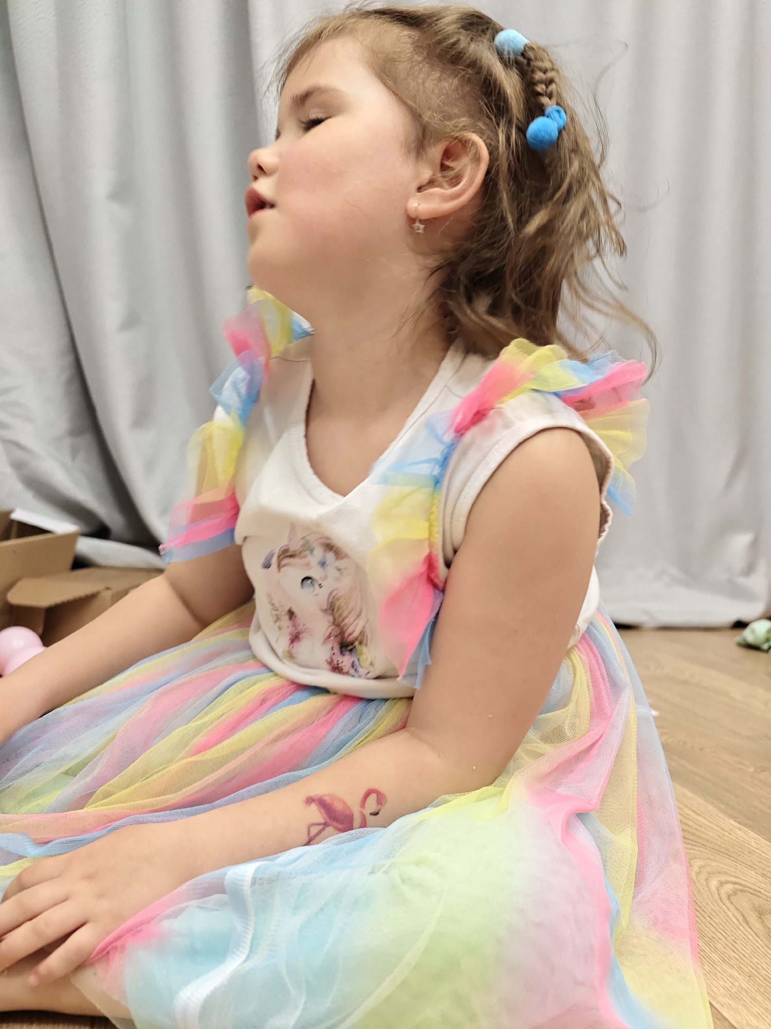 A young girl, likely a toddler or preschooler, is seated on a wooden floor in an indoor setting in Chaikovsky, Russia. Captured in profile, she faces to the left of the frame, with her eyes closed or partially closed and her head tilted slightly upwards, conveying a serene and contemplative expression.
She is wearing a colorful outfit: a white t-shirt featuring a graphic print of a white unicorn-like creature with a flowing mane, and ruffled short sleeves made of rainbow-colored tulle. Her skirt is also composed of vibrant, layered tulle in shades of pink, yellow, blue, and green. Her light brown hair is styled with two blue pom-pom hair ties securing braids, and a small silver star earring adorns her left ear. A pink flamingo temporary tattoo is visible on her right forearm.
The background consists of light-colored curtains, possibly grey or off-white. On the wooden floor near the girl, parts of a cardboard box and a pink spherical object are visible. The scene suggests a relaxed and informal moment in a home or play area. No visible text is present in the image.