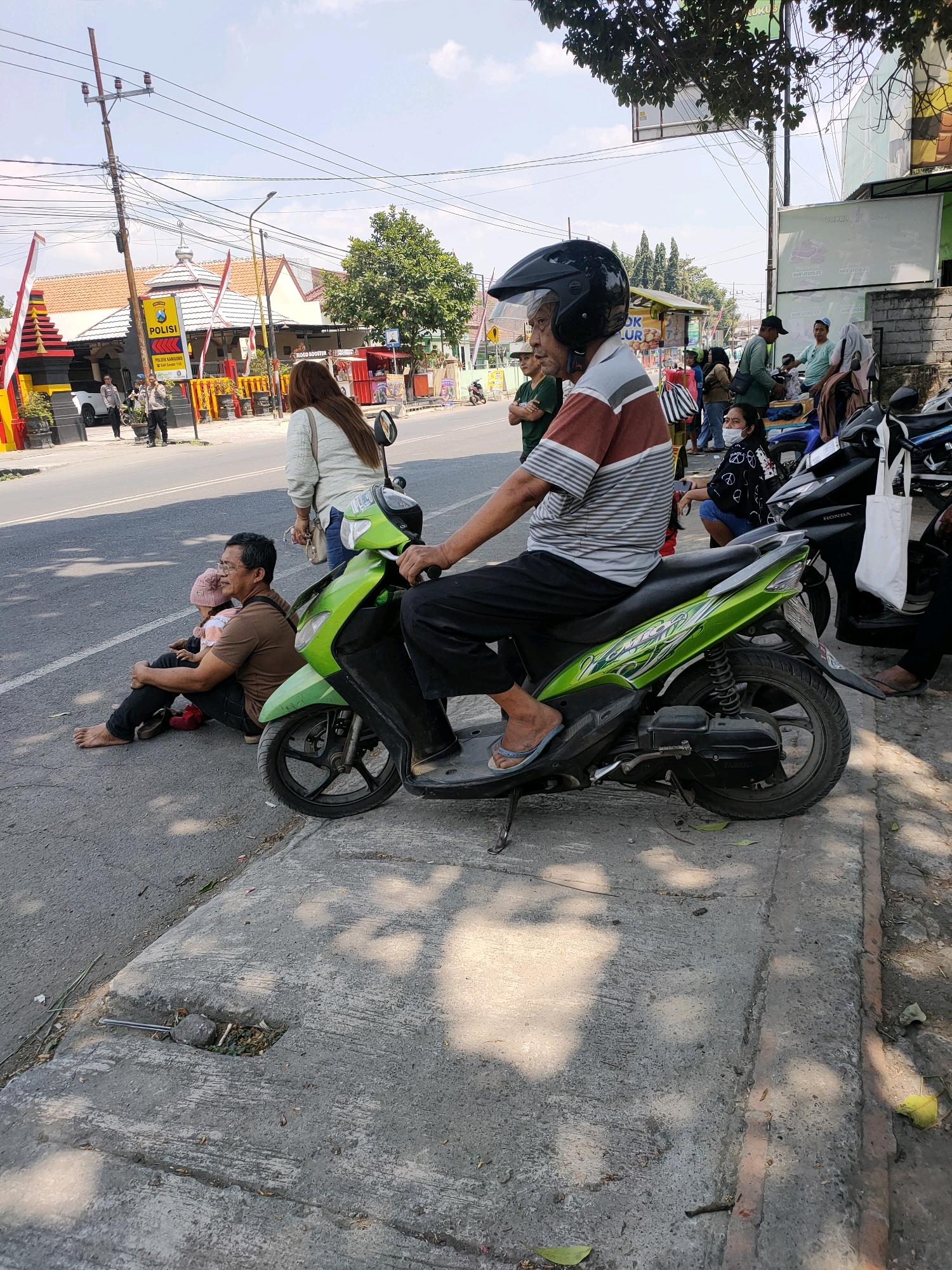 In Kanigoro, Indonesia, on a bright, sunny day, an outdoor scene unfolds along a street. In the foreground, a man wearing a helmet is seated on a green and white scooter, facing left. He is dressed in a striped shirt and dark pants. To his left, a man and a small child sit on the asphalt roadside. The man wears a brown t-shirt and dark pants, and the child is dressed in a pink hat and pink and orange clothing. A white tote bag hangs from the handlebars of a black scooter to the right of the man on the green scooter. In the background, several other people are visible, some standing and some on motorcycles, near buildings and trees. A prominent sign with the word "POLISI" indicates a police presence. Electrical wires and poles crisscross the sky, and the street is paved with asphalt, bordered by a concrete curb. The environment appears to be an urban or semi-urban setting.