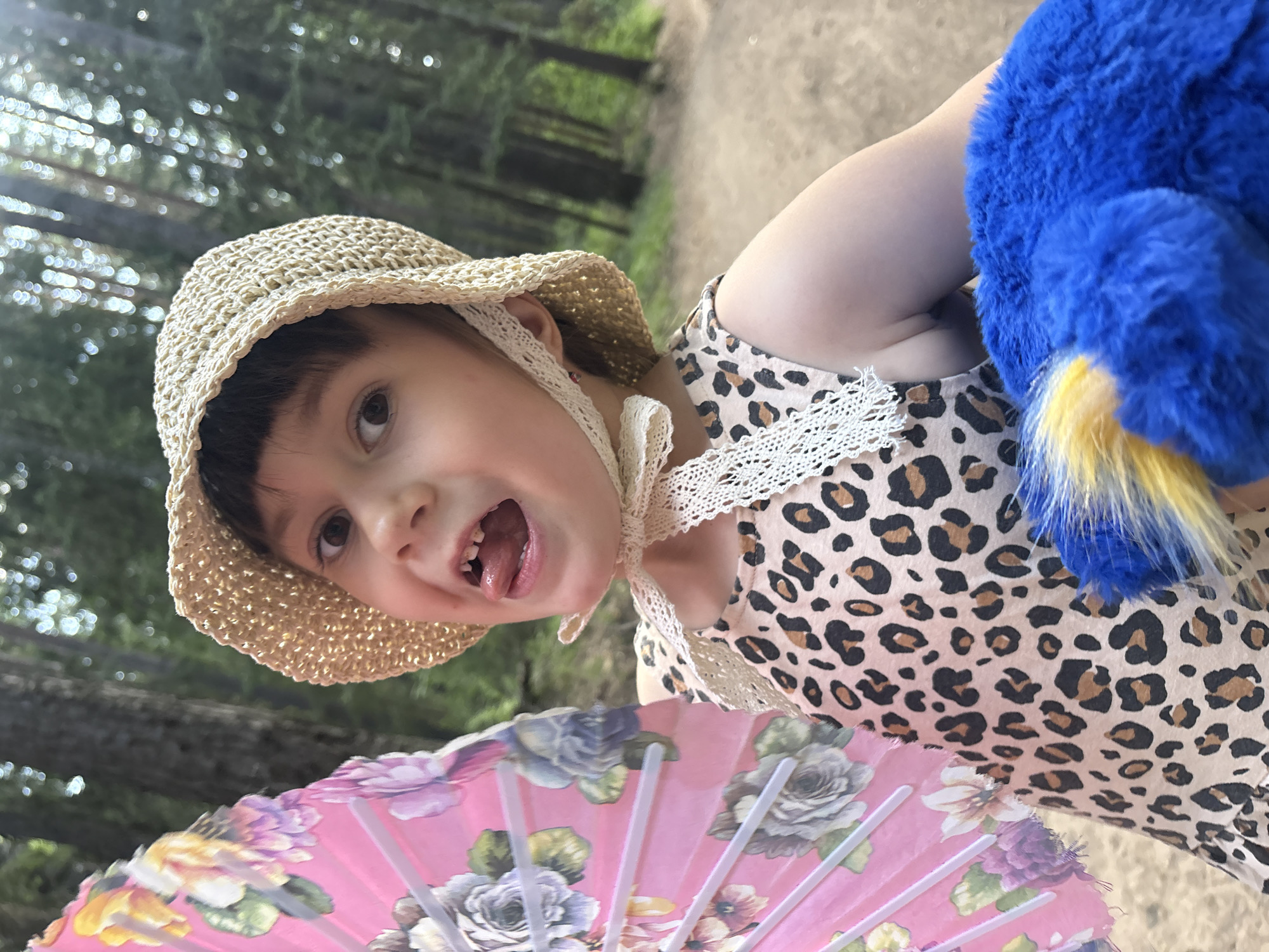 A young child in a straw hat and animal print dress is positioned at an angle facing the viewer. The child is outdoors on a path surrounded by trees. The child is holding a floral fan in front and a blue stuffed animal to the side. The child has an open mouth, revealing some missing teeth. The location is believed to be Chaikovsky, Russia.