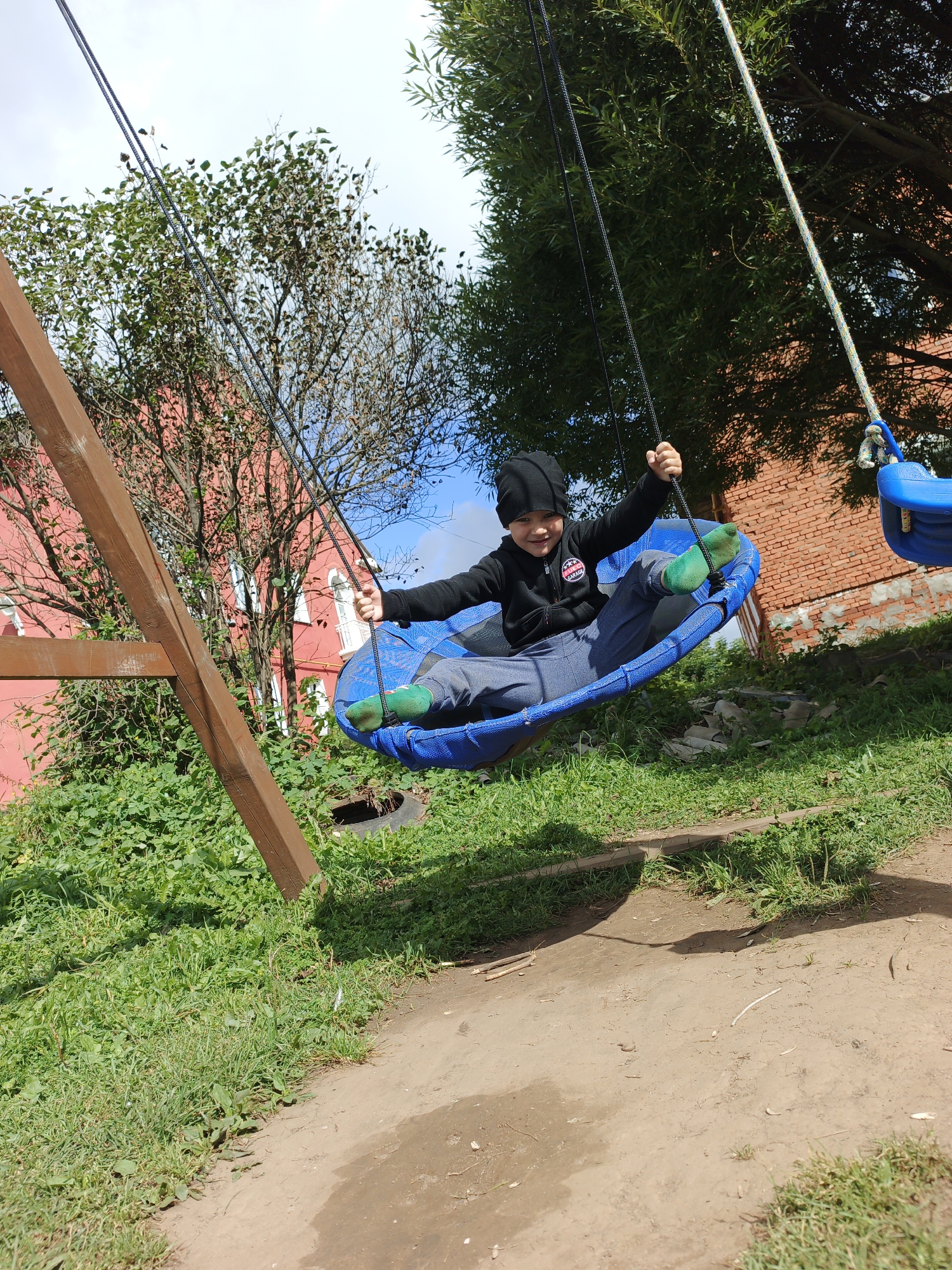 A young boy is swinging on a blue basket-shaped swing. He is wearing a black beanie, a black zip-up hoodie with a circular patch on the left breast, grey pants, and green socks. The boy is smiling and looking at the camera with his arms outstretched. The swing is suspended by ropes from a wooden swing set frame. In the background, there is a red brick building with windows and a red wall with some trees in front of it. The ground is covered in grass and dirt. The sky is blue with some clouds. The location is a park in Medvedyovka, Russia.