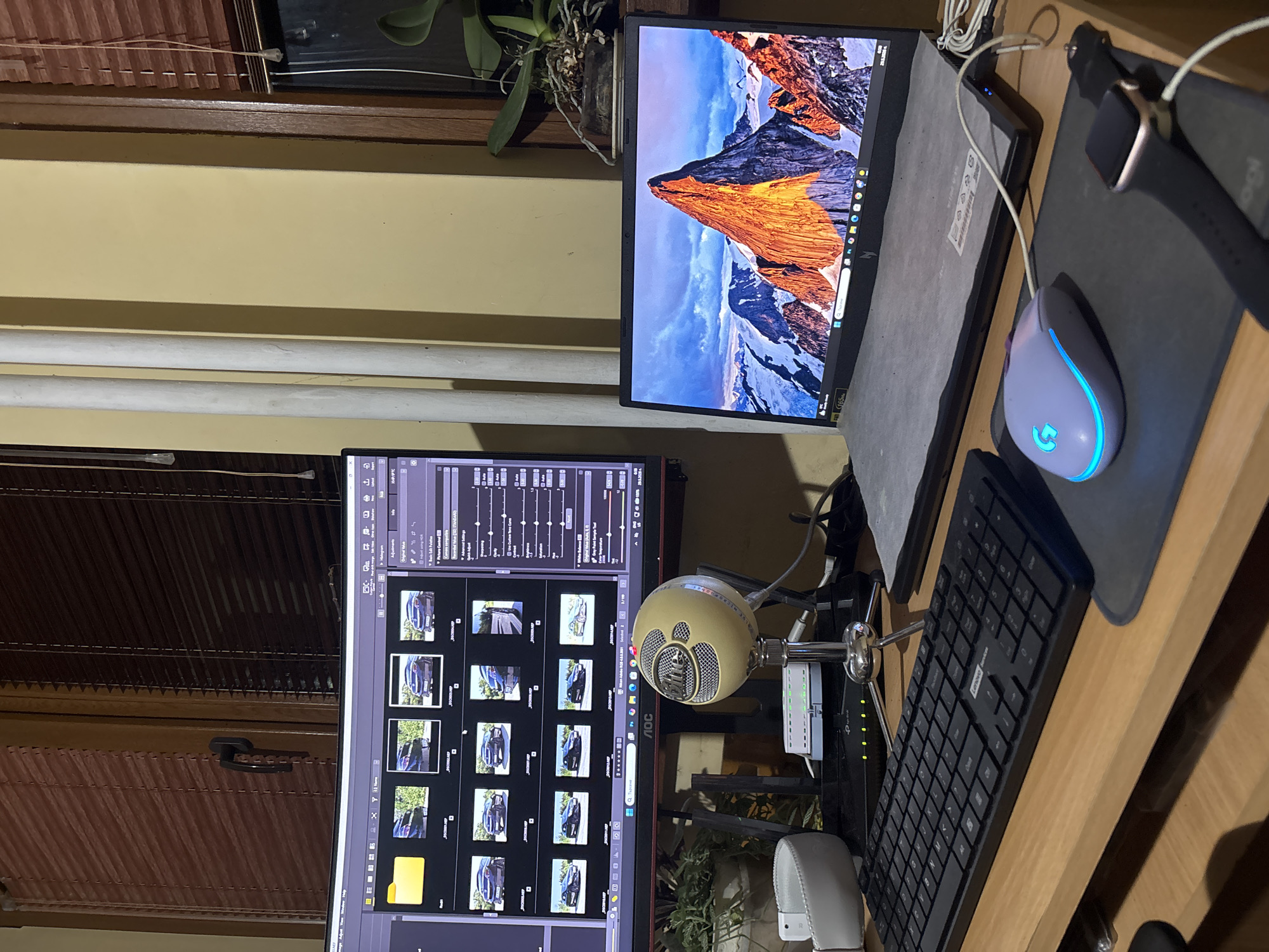 The image presents a desk cluttered with technology. Two monitors are visible: one displays a mountain range with snow and a vibrant orange peak, while the other features what appears to be photo editing software with numerous images of a black car. A Blue Yeti microphone sits in front of the lower monitor, along with a black Lenovo keyboard and a white Logitech G mouse on a black mousepad. An Apple Watch rests on the mousepad. A TP-Link network switch is positioned near the microphone. The background includes a wall with blinds, houseplants, and what appears to be a window. The location is indeterminable from the image, so including the location in Lovech, Bulgaria would be inaccurate.