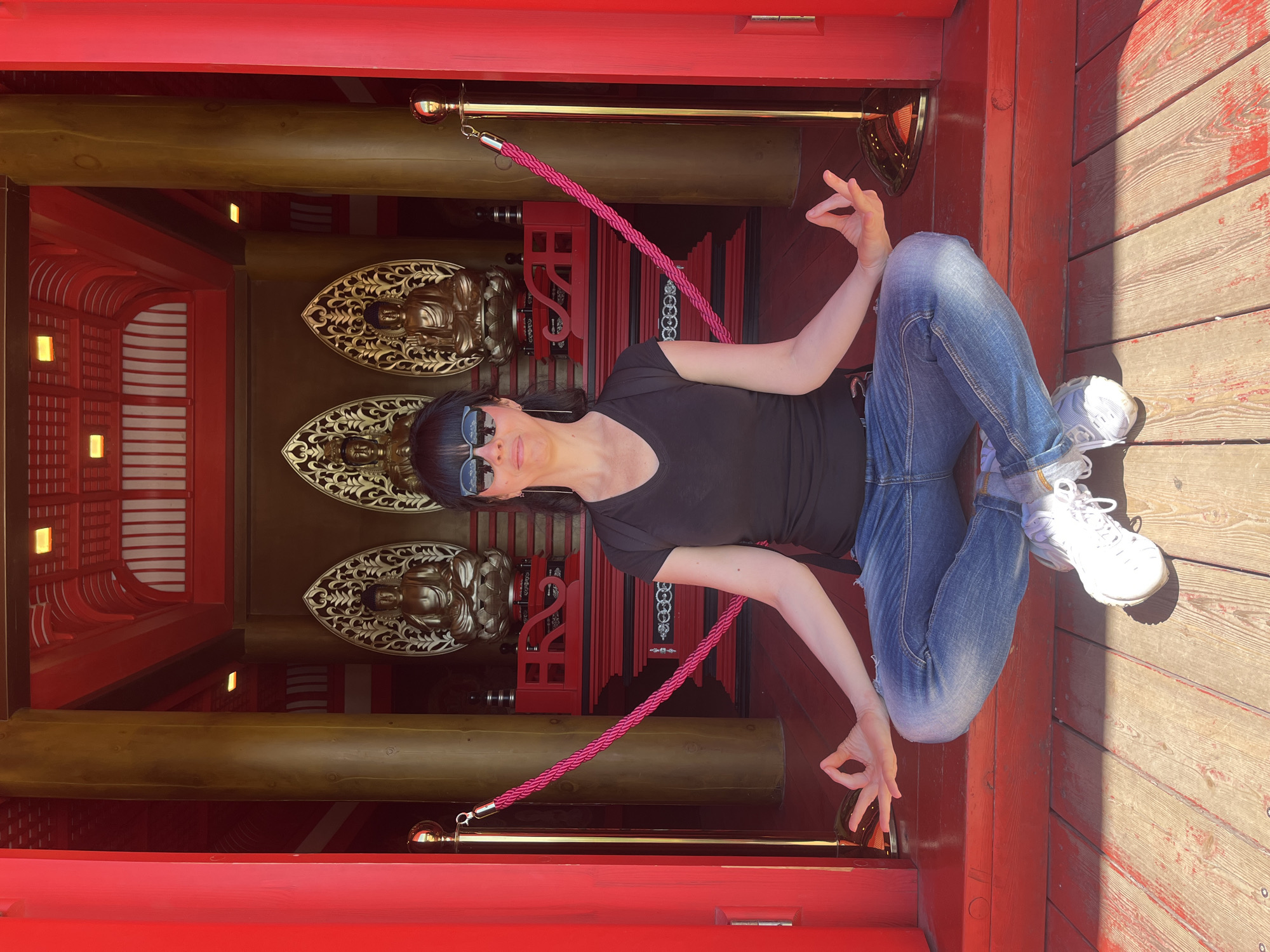 A full-shot image, rotated ninety degrees counter-clockwise, depicts a woman sitting cross-legged in a contemplative or meditative pose on a reddish-brown wooden floor. She is wearing a black v-neck t-shirt, blue denim jeans, and white athletic shoes, with dark sunglasses obscuring her eyes. Her hands are positioned in mudras, with the thumb and forefinger forming a circle.
The background features rich, reddish-orange architectural structures, possibly wooden, forming an alcove or interior space. Within this alcove, three ornate golden reliefs of seated Buddha figures are vertically aligned on a darker surface, each surrounded by intricate golden filigree. Thick, dark wooden beams span across the upper and middle sections of the background. A bright pink braided rope barrier, held by golden stanchions, is visible in the mid-ground, extending diagonally.
The scene is brightly illuminated by sunlight, casting distinct shadows on the wooden surfaces, suggesting a clear, sunny day. The architectural style, Buddha figures, and the woman's meditative posture create an atmosphere reminiscent of an East Asian temple or cultural center. The setting is in Krasnodar, Russia. No text is visible in the image.