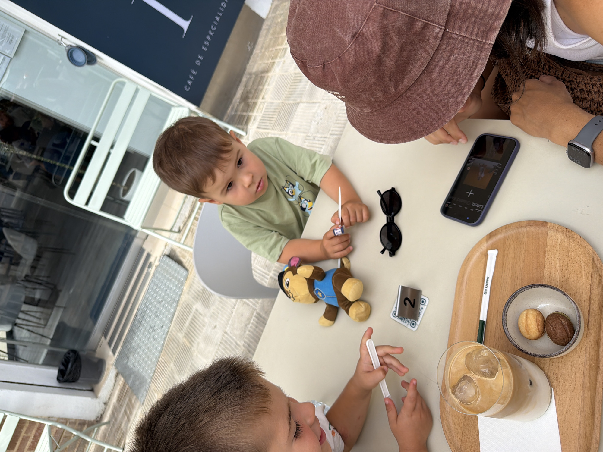 Two young boys and an adult are seated at a table outdoors, likely at a cafe. One boy, wearing a light green t-shirt with cartoon characters, is looking down at the table, holding a small white object. He is seated in the middle of the frame. To his left, another boy, partially visible, is also looking down and holding a white object. In front of the middle boy is a stuffed toy, resembling a dog wearing a blue vest and hat. To the right of the stuffed toy is a small metal sign with the number "2" and a QR code. Further to the right, a person wearing a burgundy bucket hat is looking down at a smartphone. On the table, to the left, is a glass of iced coffee with a paper straw, and a small bowl containing two pastries. The background shows outdoor seating, glass doors, and some foliage. The overall scene suggests a casual outing.