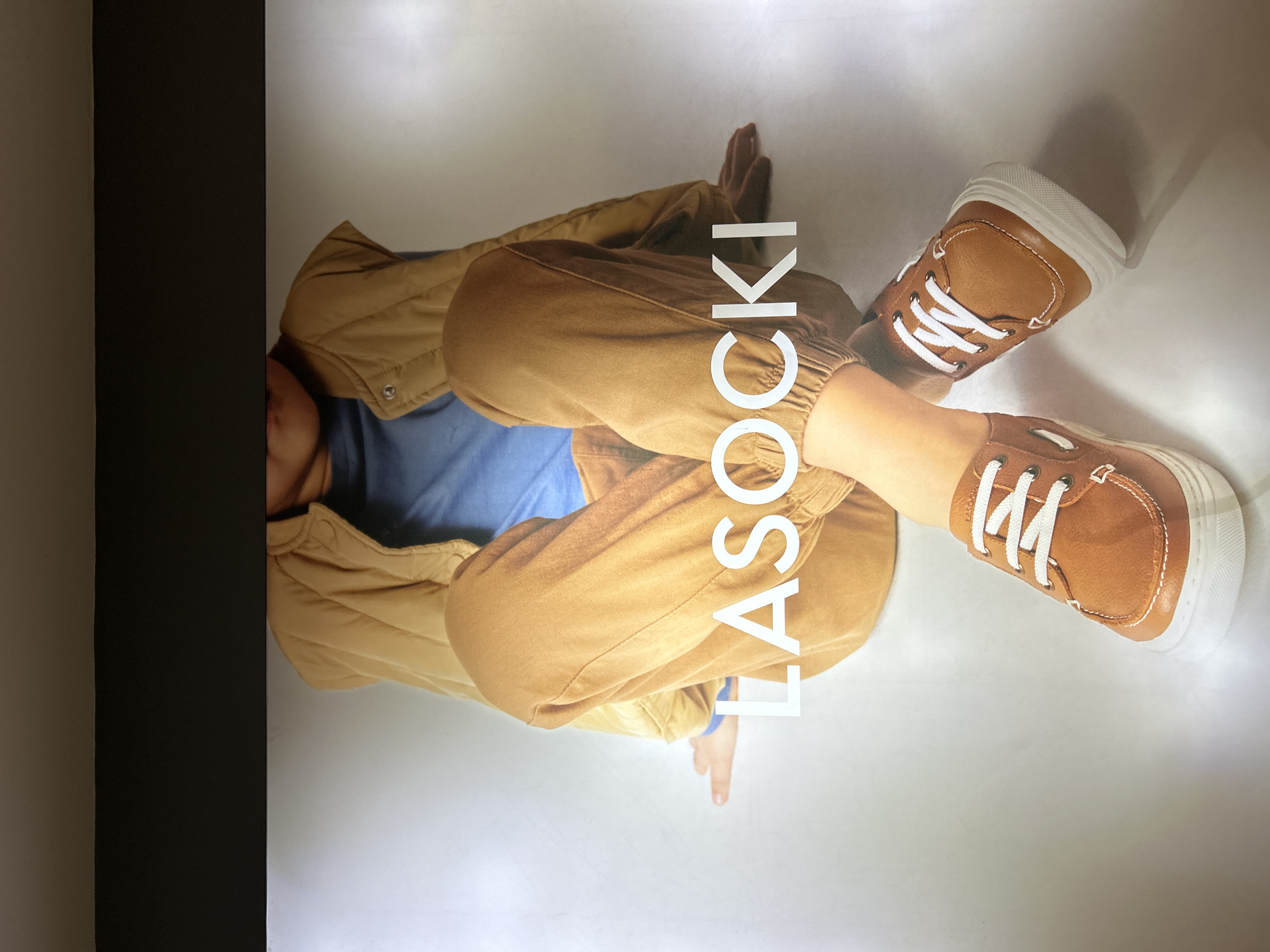 The image shows a child sitting on a white surface. The child is wearing a blue shirt, tan pants, a tan jacket, and tan shoes with white laces and white soles. The word "LASOCKI" is written in white letters on the image. The image is likely a promotional advertisement for LASOCKI brand shoes. There is no discernible information to indicate the photo was taken in Plovdiv, Bulgaria. The photo appears to be taken indoors, next to a dark wall.